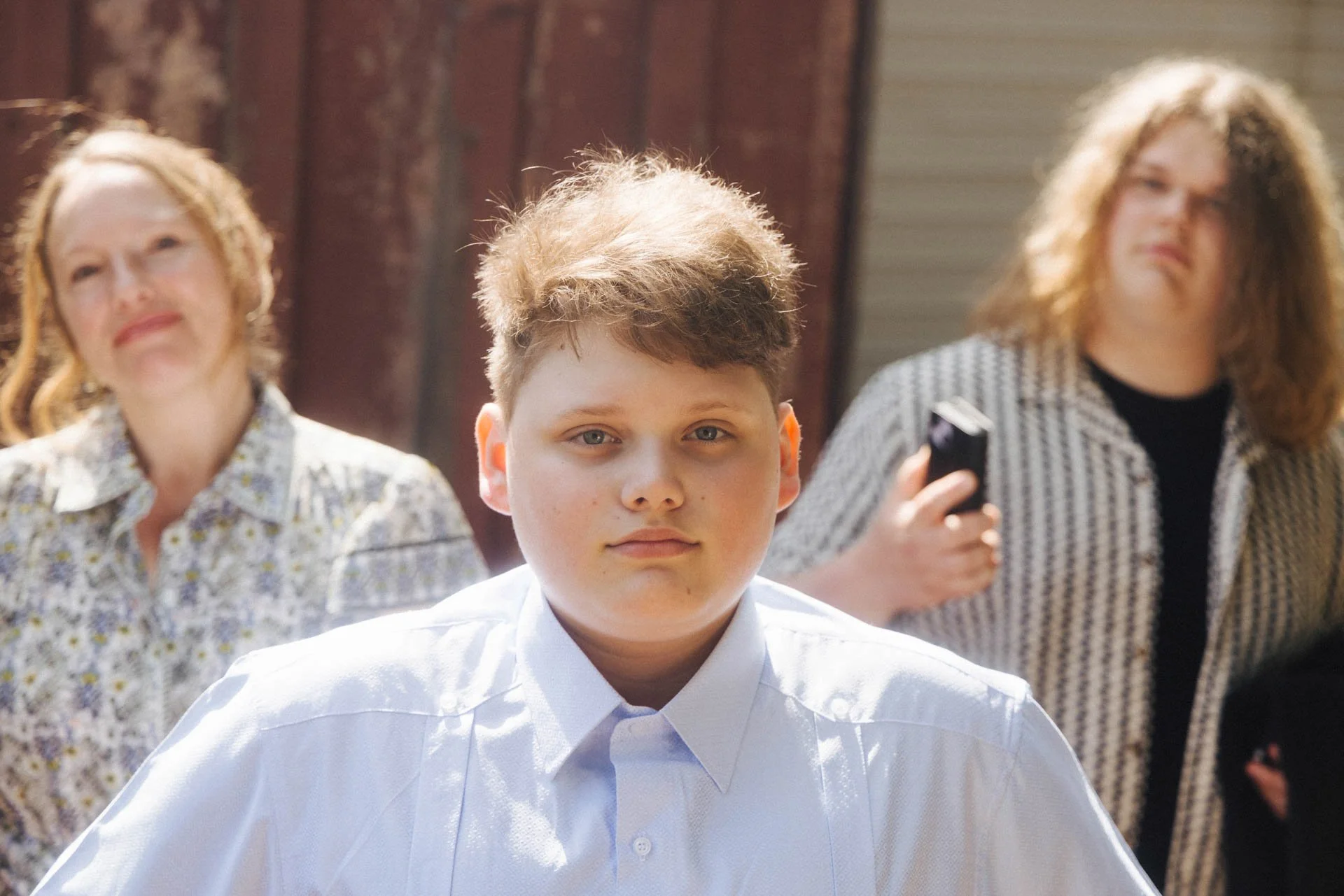 Young boy with Blond hair and a white shirt in the foreground, with two women blurred in background, one holding a phone, outside with wooden and metal structures.