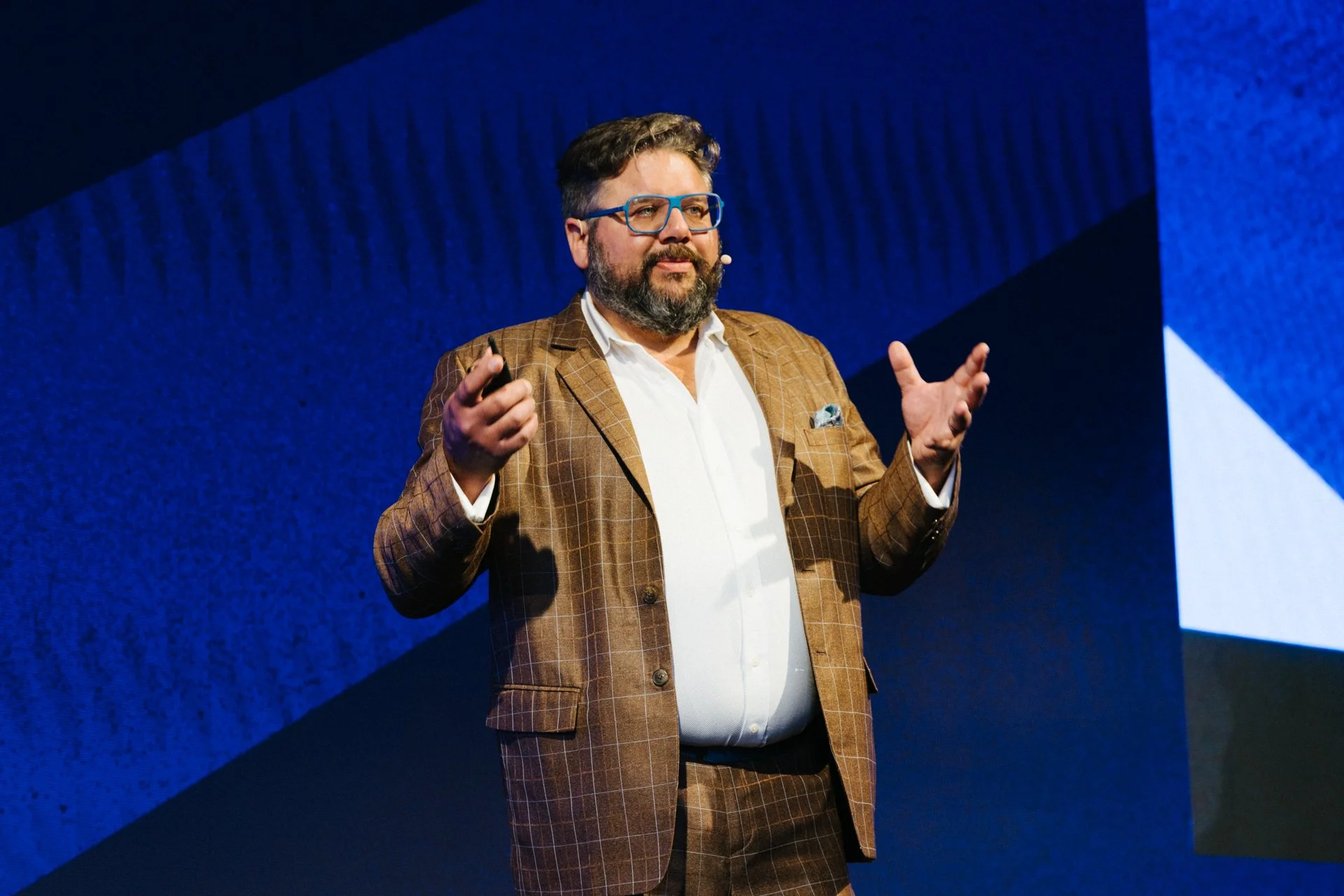 Man wearing glasses and a brown checkered suit giving a presentation on stage with a blue background.