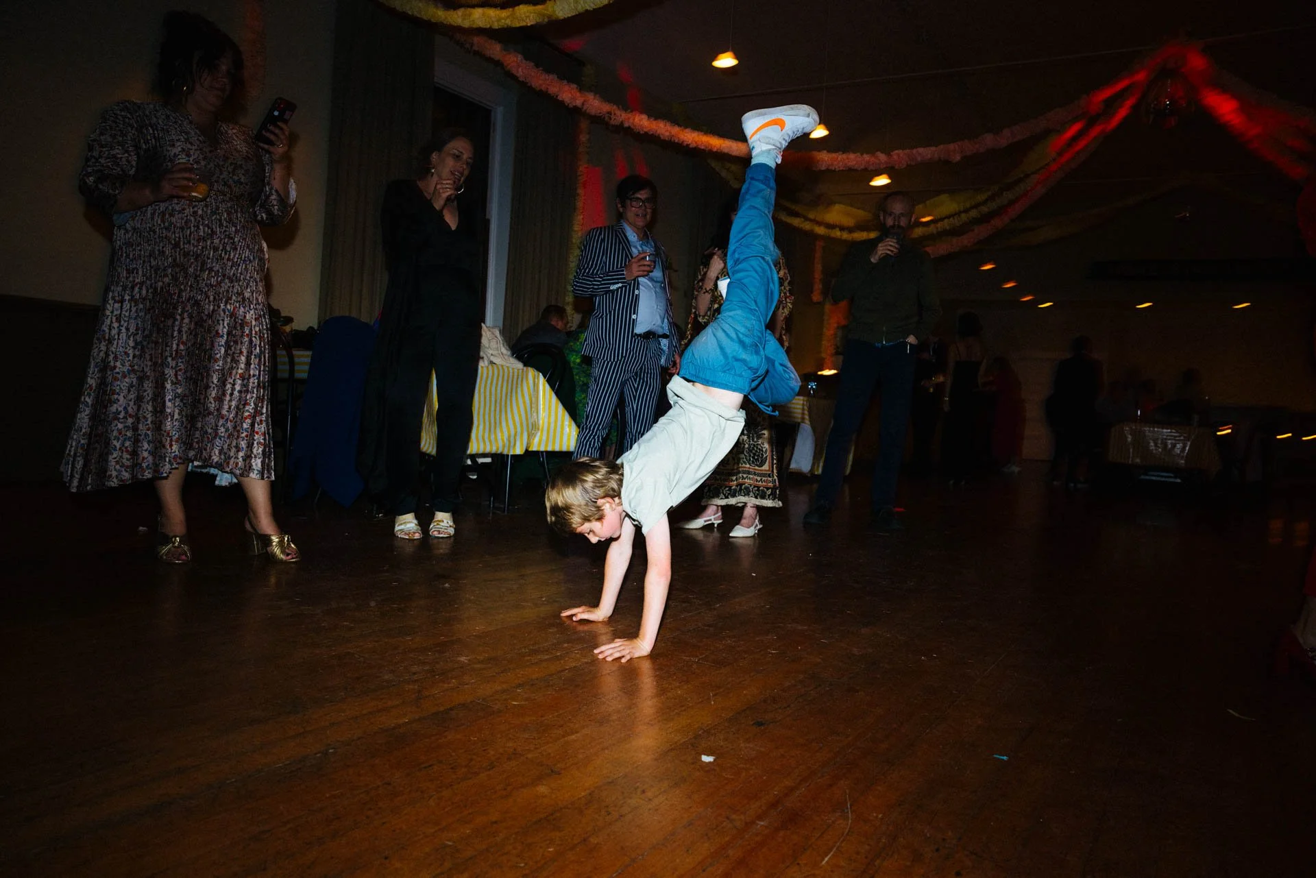 A young boy performing a handstand on a wooden dance floor at an indoor party, with several adults watching and mingling in the background, decorated with streamers.