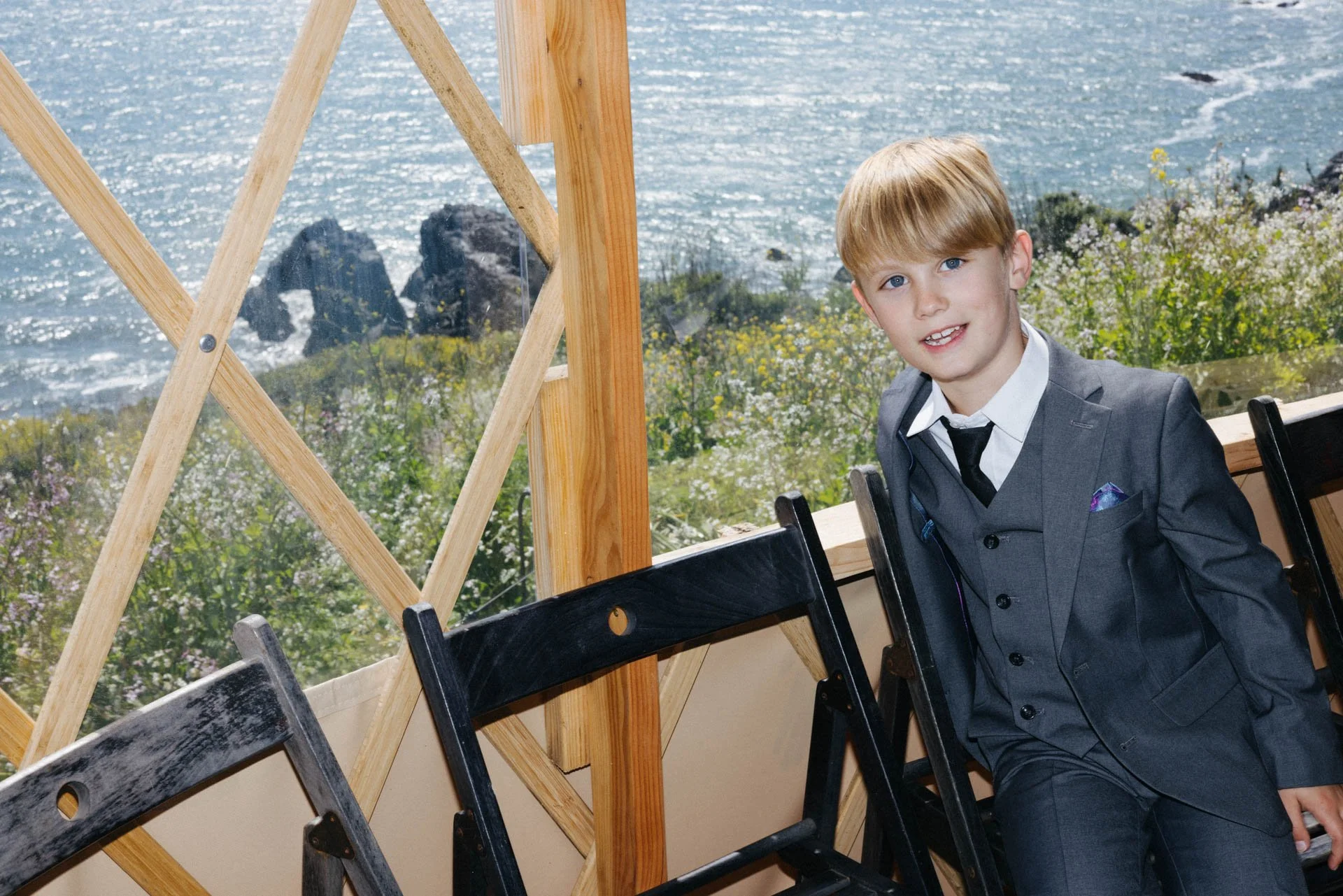 A young boy in a gray suit with a black tie sitting indoors near a large window with a view of the ocean and rocky coastline, with wooden beams and black chairs in the foreground.
