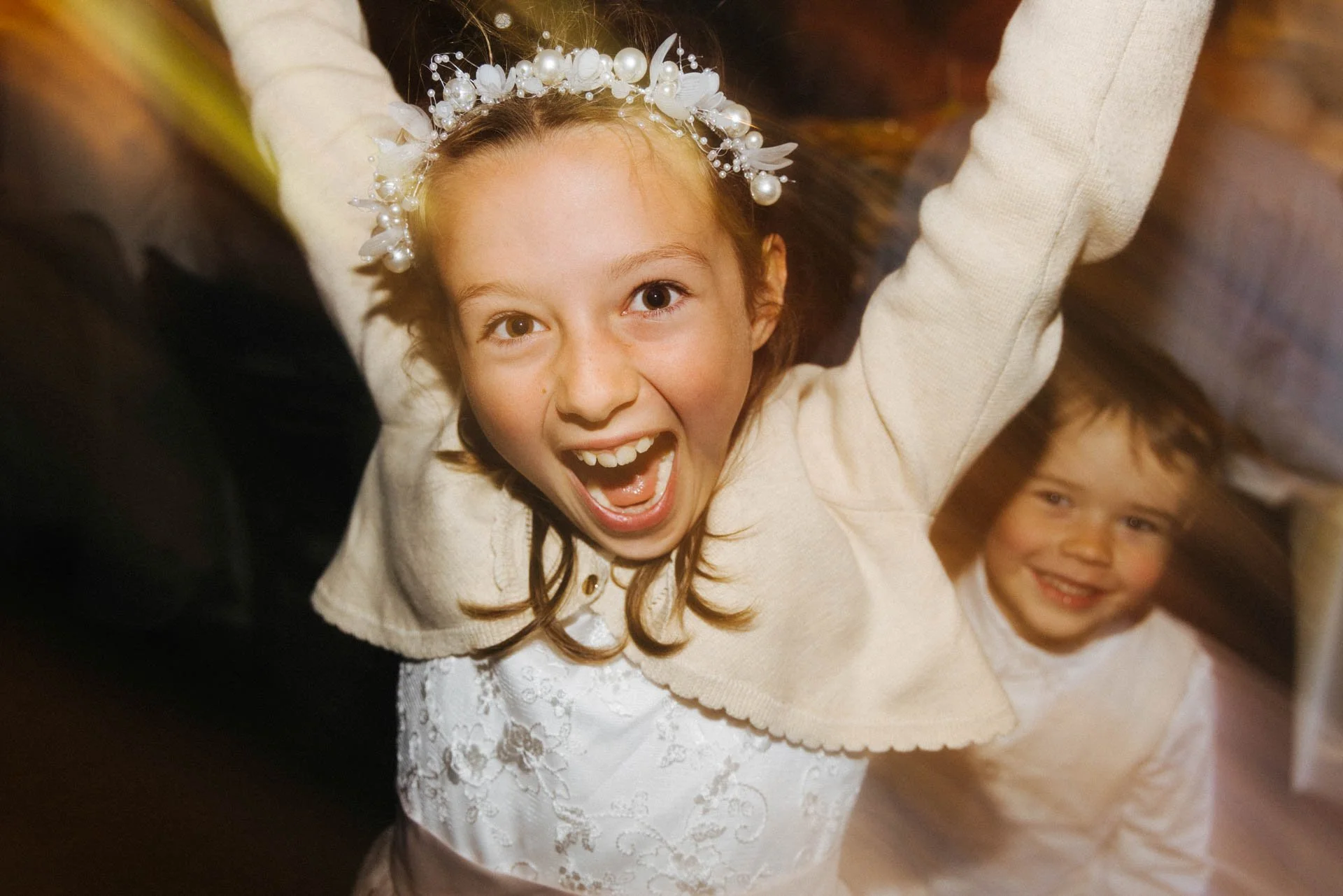 Two young girls wearing white dresses, celebrating and smiling, with one girl wearing a pearl and floral headband, in a festive setting.