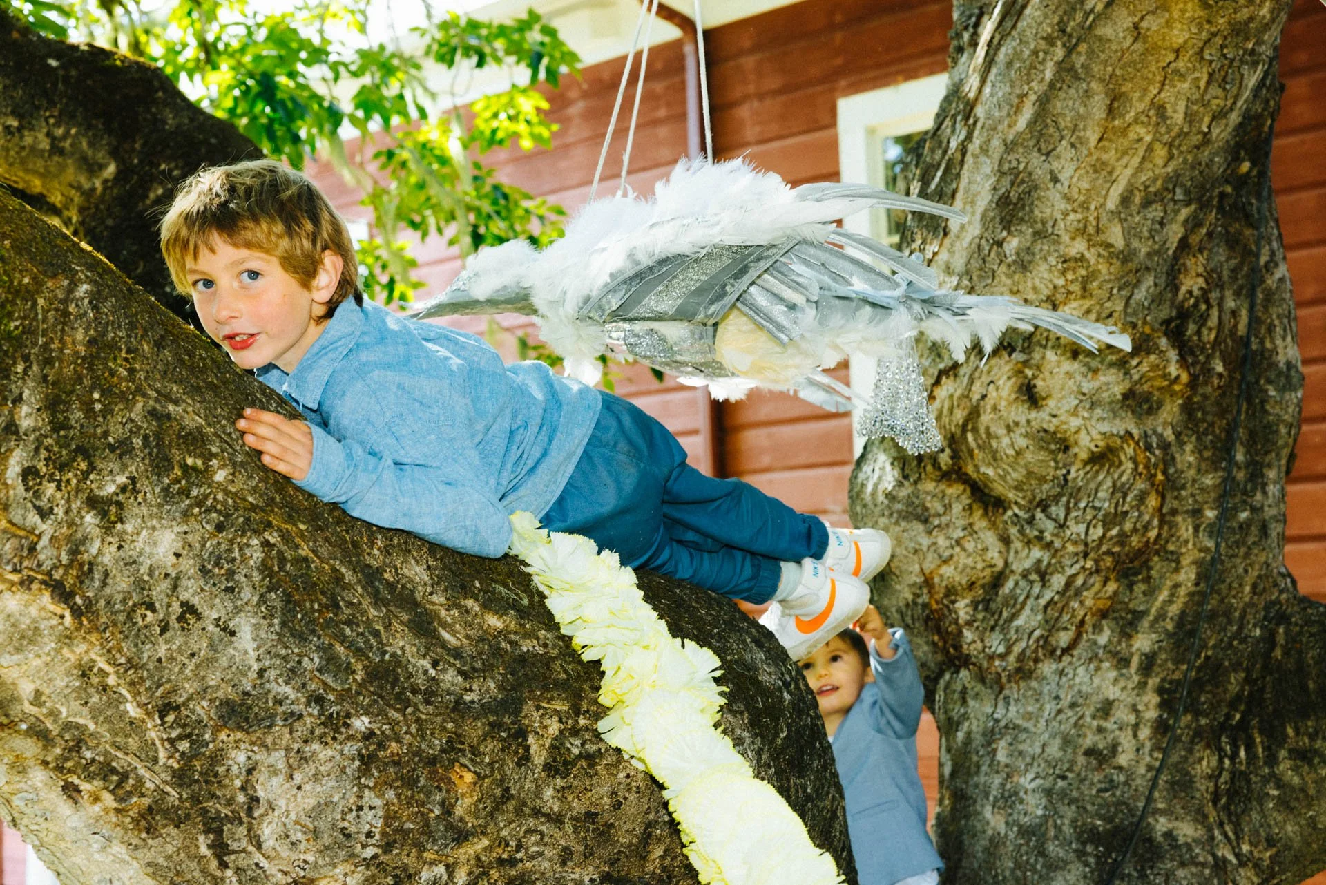 Child 1 wearing a blue shirt and jeans lying on a tree branch, looking towards camera, with a hanging decorative bird made of feathers and metallic parts. Child 2 in the background, pointing upwards.