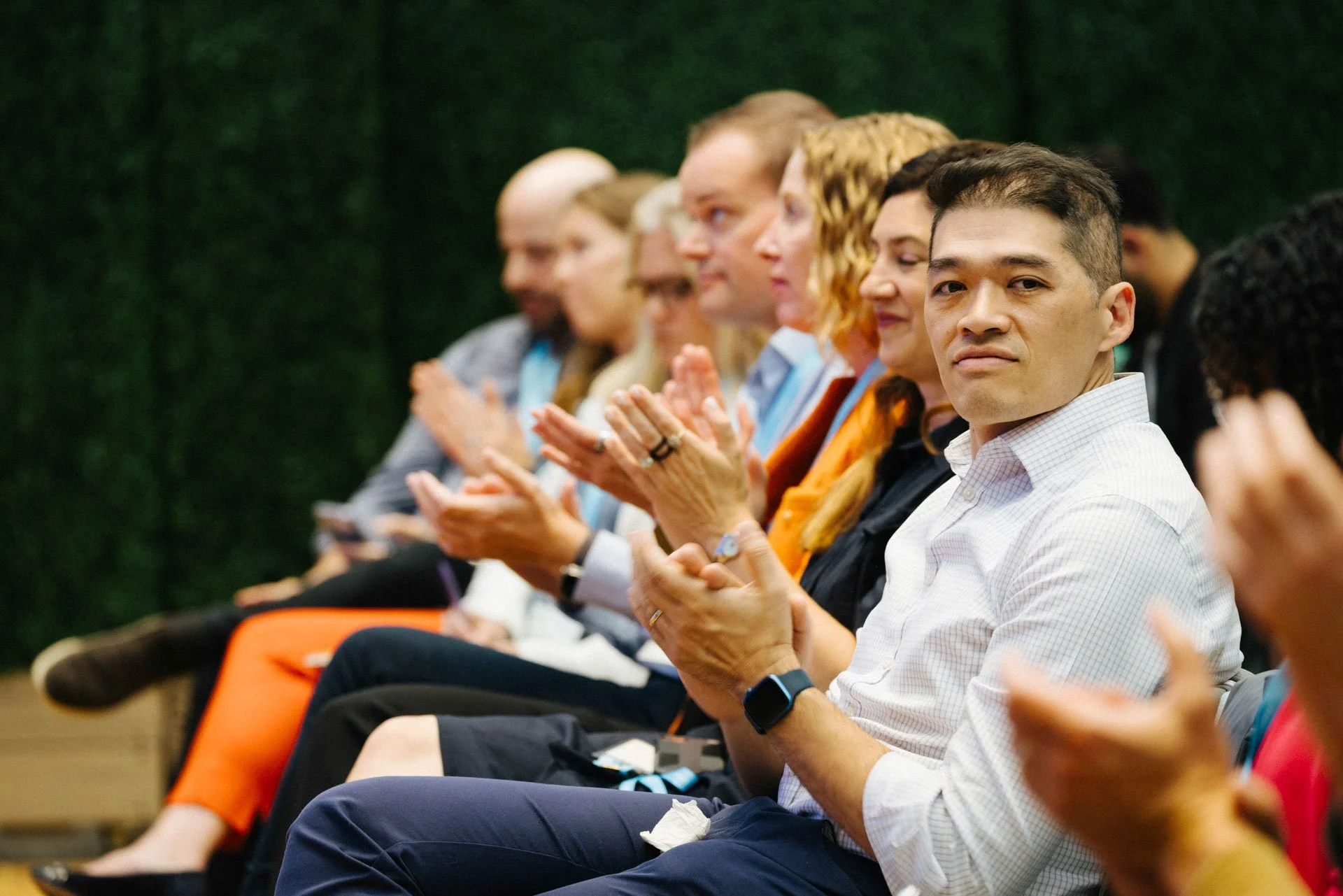 Audience seated in a row at an indoor event, clapping and paying attention to a presentation.