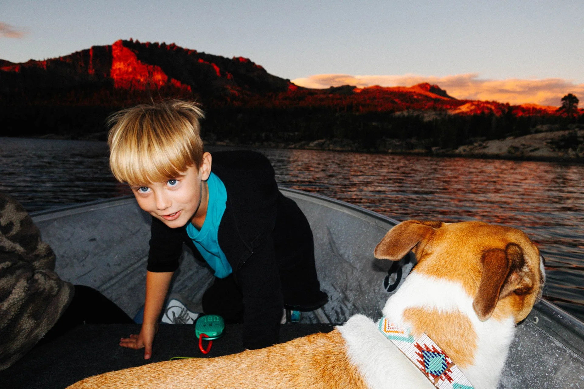 A young boy with blond hair and blue eyes in a black hoodie and turquoise shirt in a boat with a brown and white dog. The boy is crawling on the boat with water and a mountain landscape at sunset in the background.