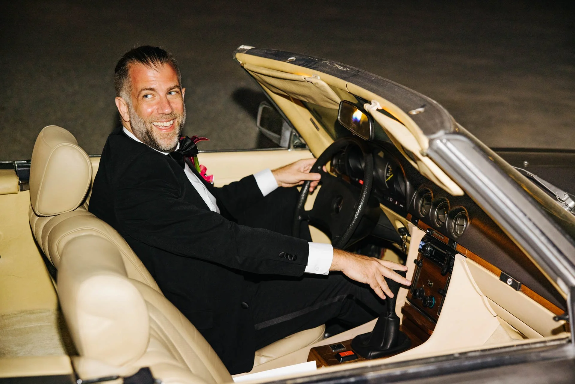 A man in formal attire sitting in the driver's seat of a vintage convertible car, smiling and looking back over his shoulder.