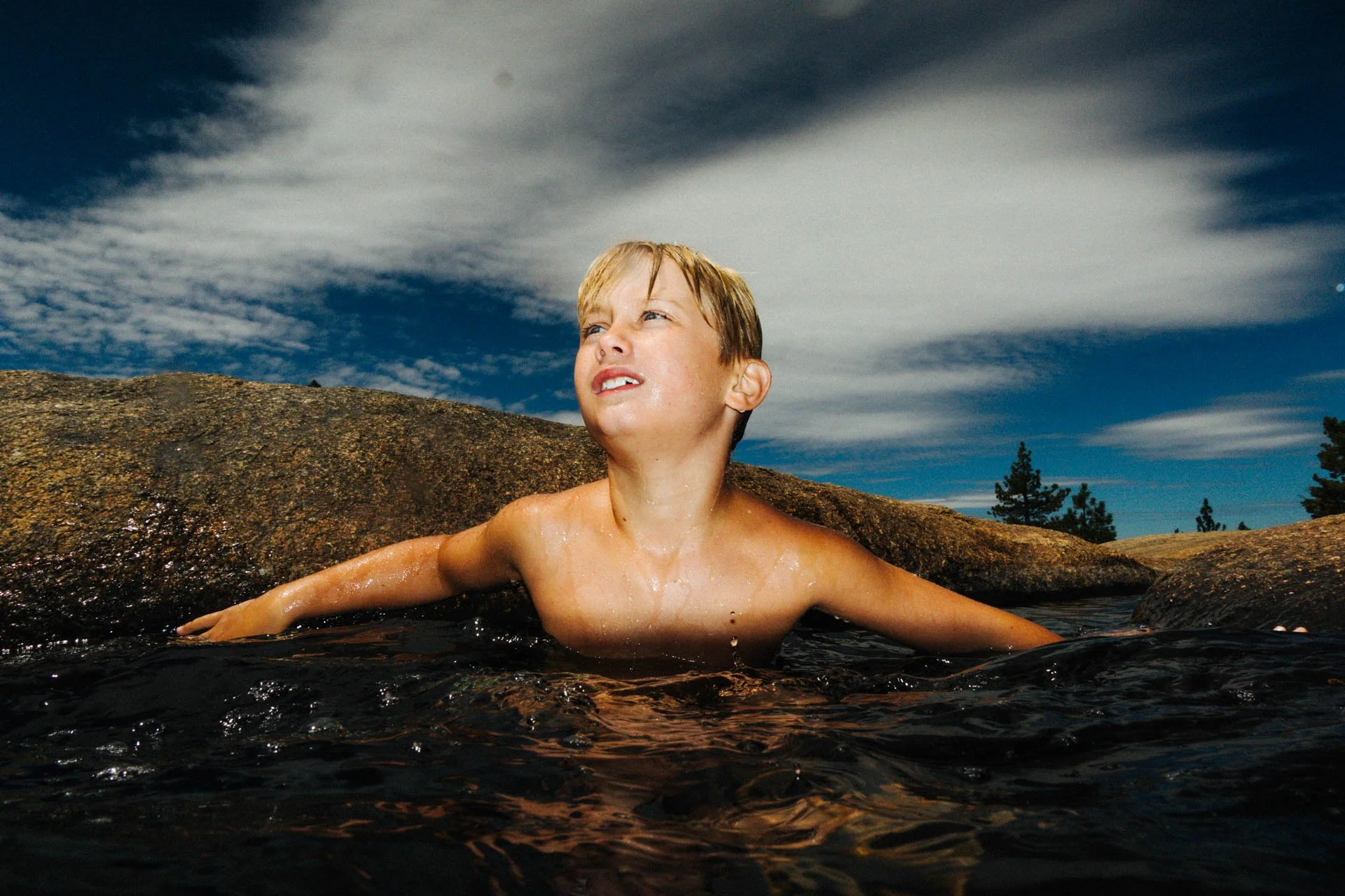 Young boy swimming in a natural outdoor pool with rocks, looking up at the sky.