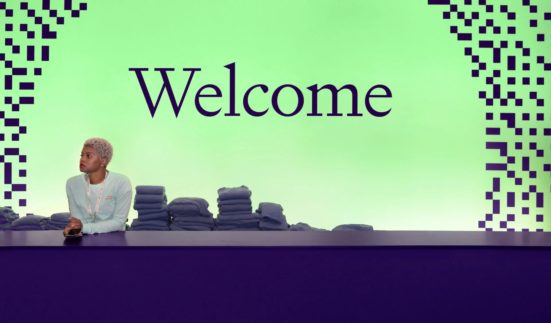 A woman with short, platinum blonde hair sitting at a registration or information counter at an event, with multiple stacked folded clothes, and a large screen behind her displaying the word 'Welcome' on a green background with black and white pixela