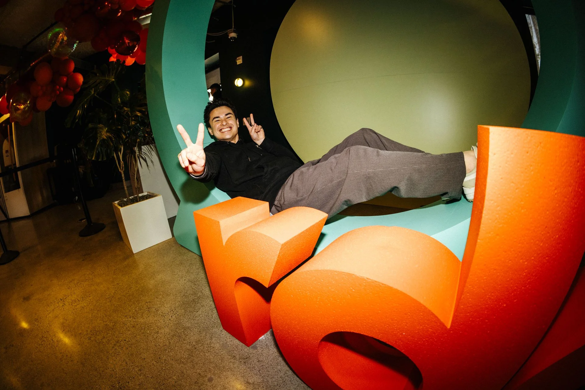 A young man is lying inside a large, colorful, curved chair shaped like the letter 'L', making peace signs with both hands, smiling widely at the camera.