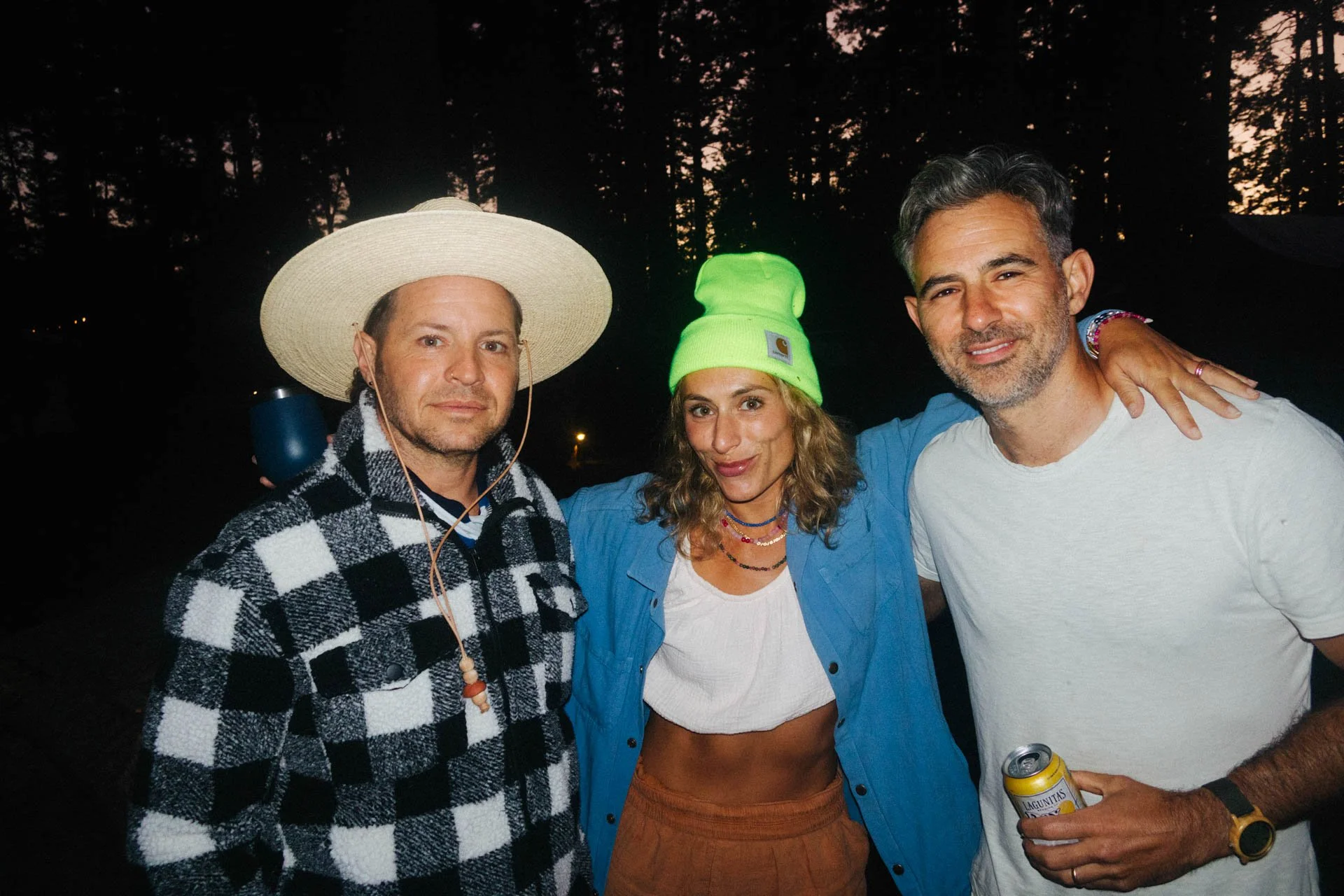 Three people standing outdoors at dusk. The person on the left is wearing a large straw hat, a black and white checkered jacket, and headphones. The person in the middle is wearing a bright green beanie, a blue shirt, and a white crop top. The person
