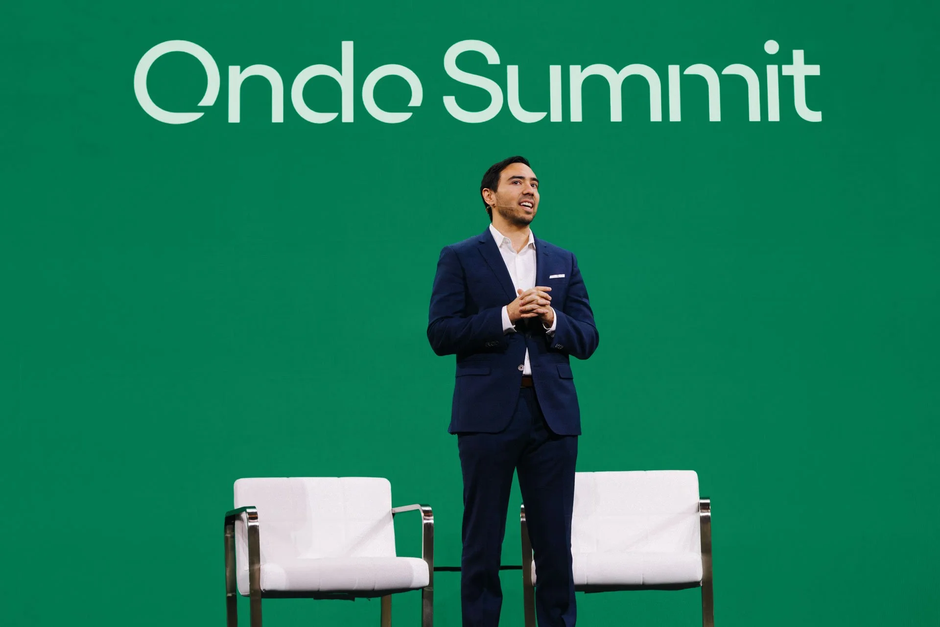 A man in a navy suit speaking on stage at an event with green backdrop that reads 'Onde Summit', with two white chairs behind him.