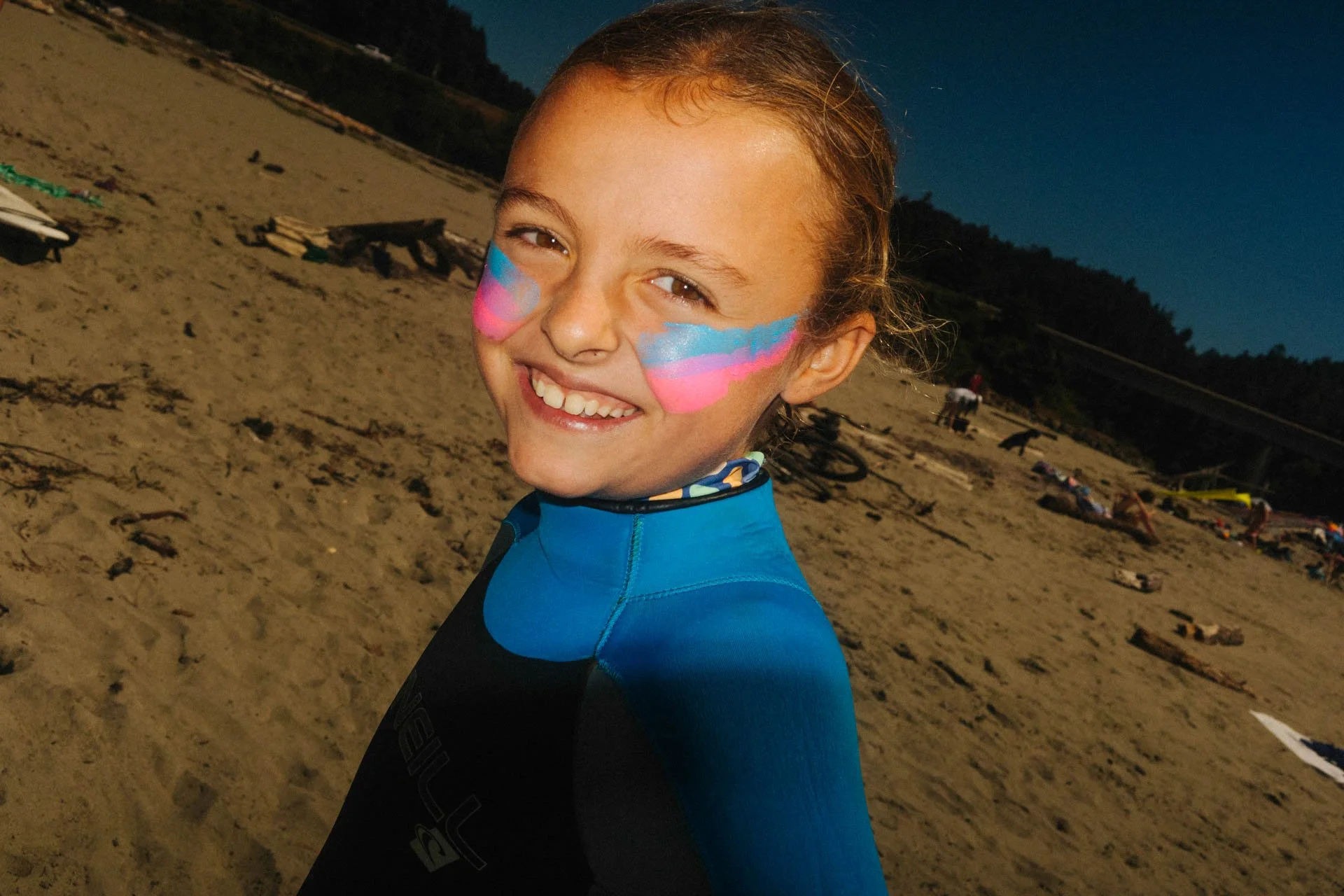 Smiling young girl with colorful face paint at the beach during early evening, wearing a blue swim shirt, with beach and some people and bicycles in the background.
