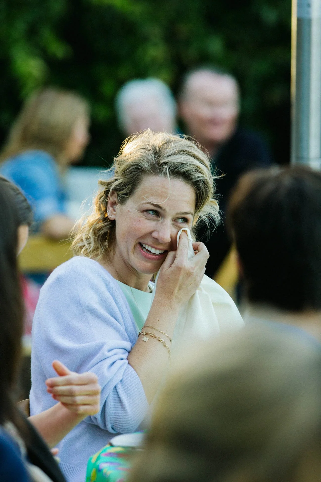 A woman with blonde curly hair smiling and wiping away tears with a tissue at an outdoor gathering, surrounded by other people.