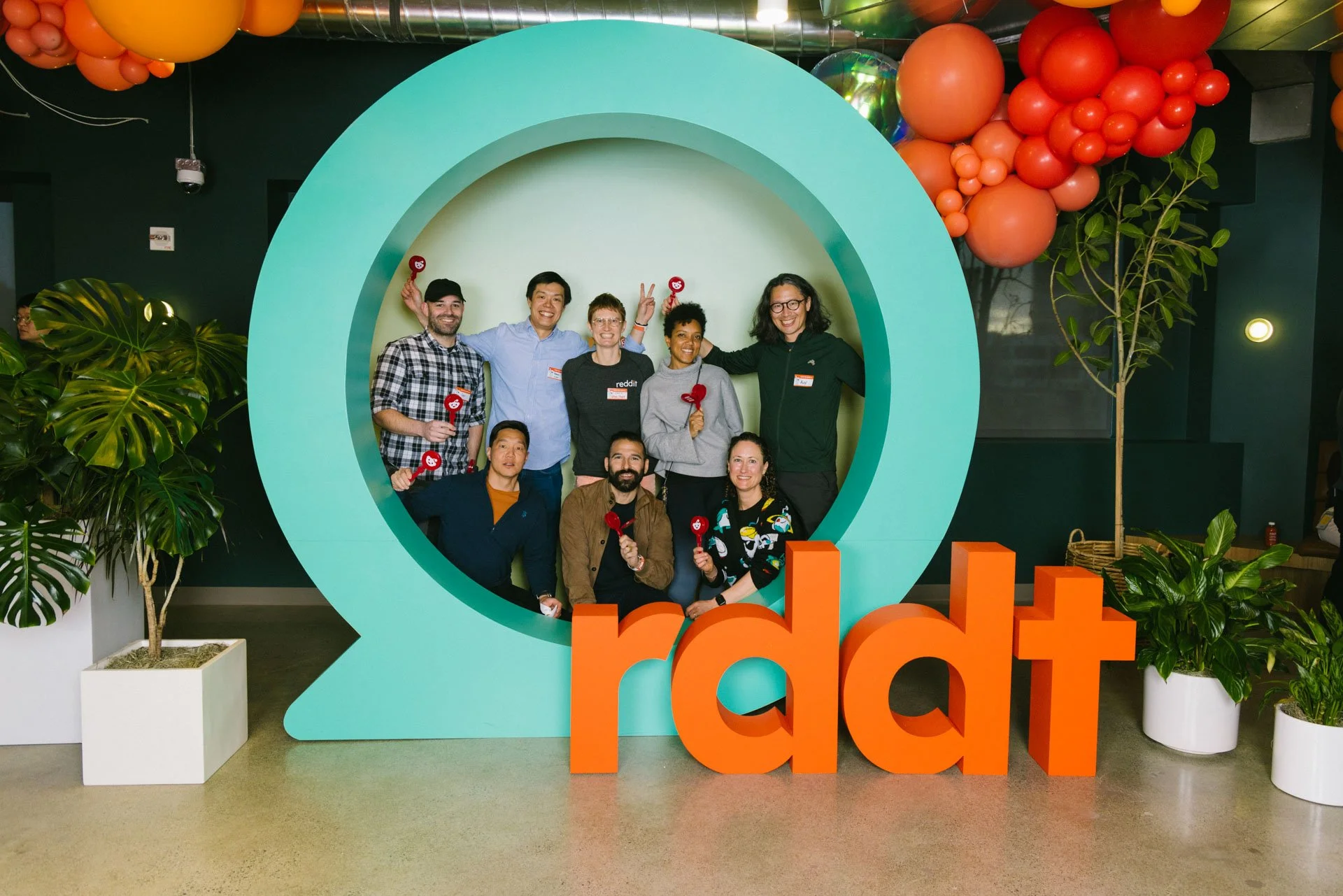 Group of nine people posing inside a large Reddit logo, with the word 'reddit' in large orange letters at the base, surrounded by plants and orange balloons.