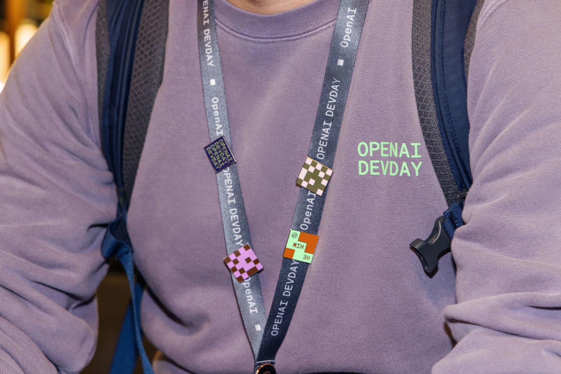 Person wearing a purple hoodie with green text reading 'OPENAI DEV DAY,' with a black lanyard around their neck featuring badges and pins, and a backpack strap over their shoulder.