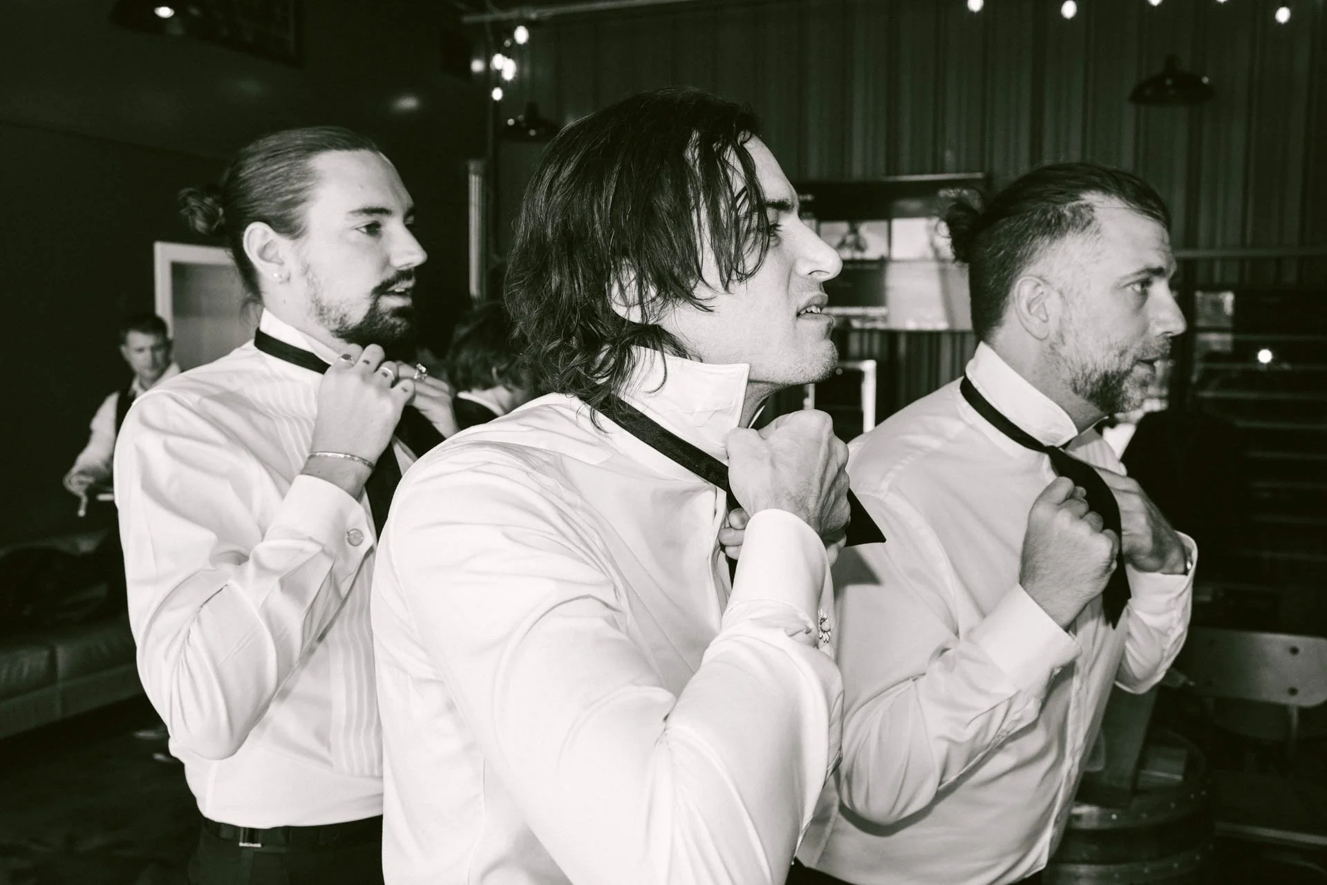 Three men adjusting their bow ties in a line, wearing white shirts, in a setting that appears to be a backstage area or dressing room.