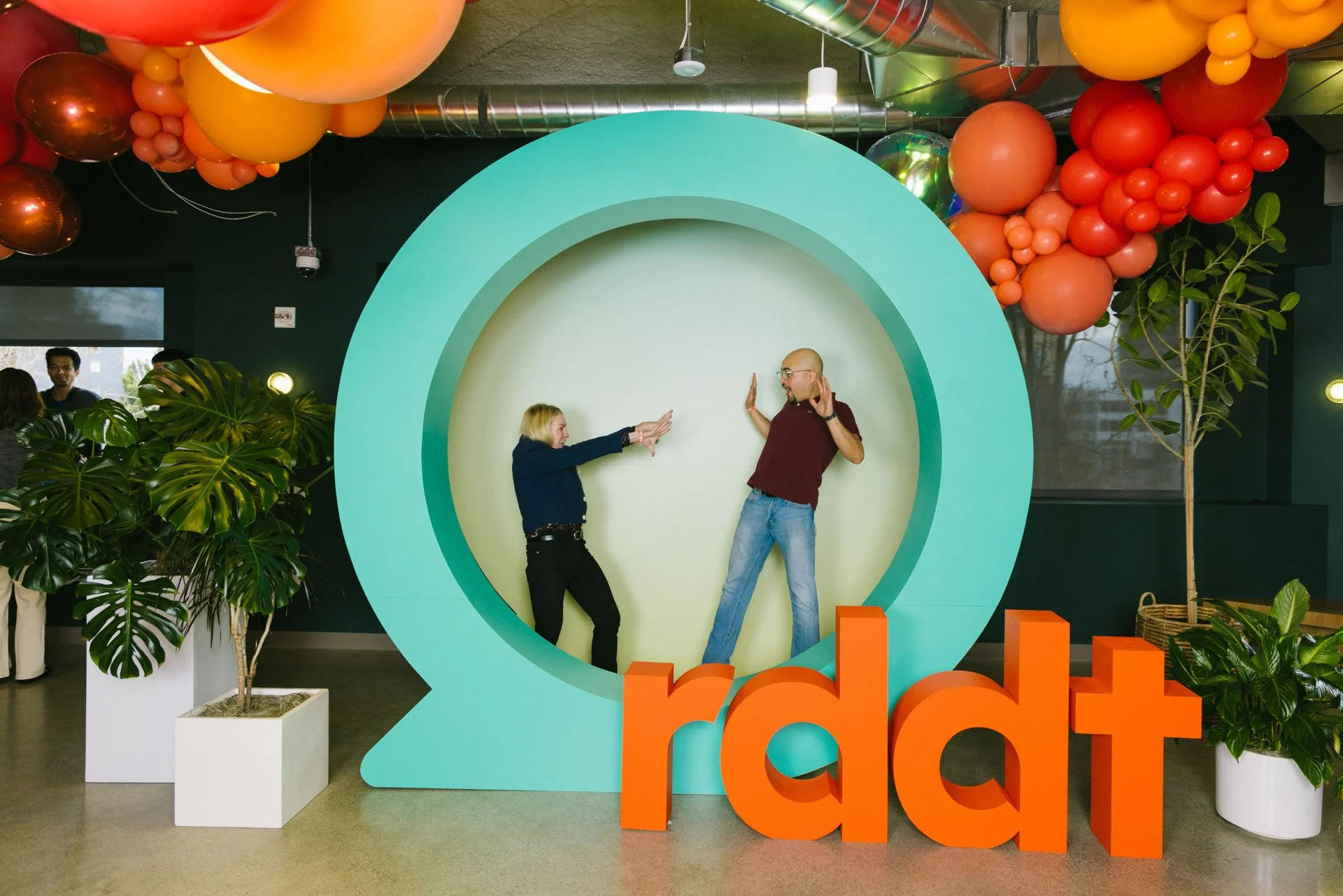 Two people posing playfully in front of a large Reddit logo at an indoor event space decorated with colorful balloons and plants.