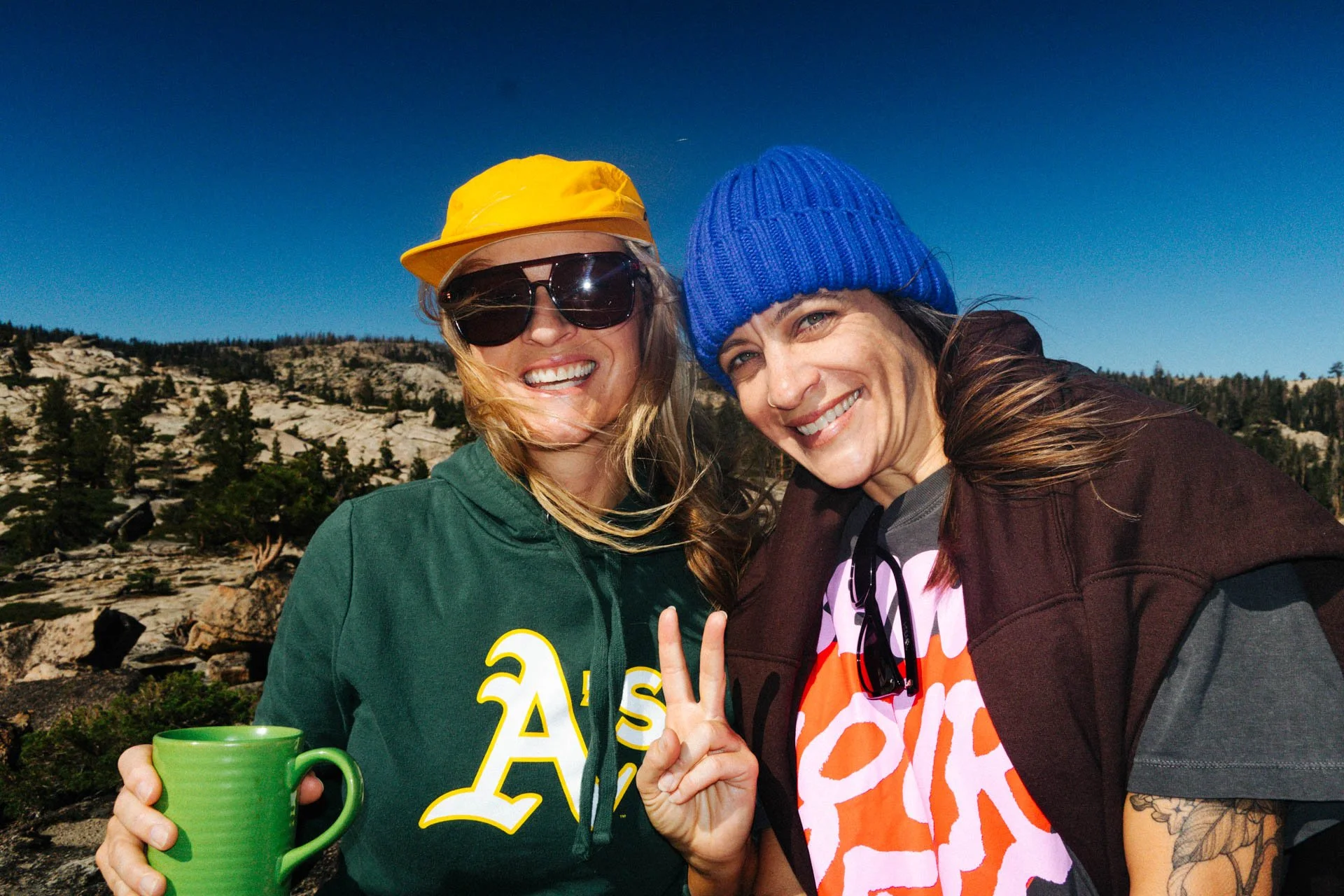 Two smiling women outdoors, one wearing a yellow cap and sunglasses, holding a green mug, and the other wearing a blue beanie, with a mountain landscape and clear blue sky in the background.