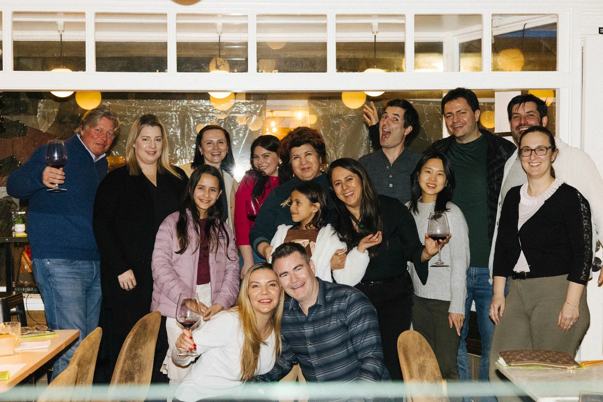 Group of people celebrating indoors, some holding glasses of wine, smiling, and posing for the photo.