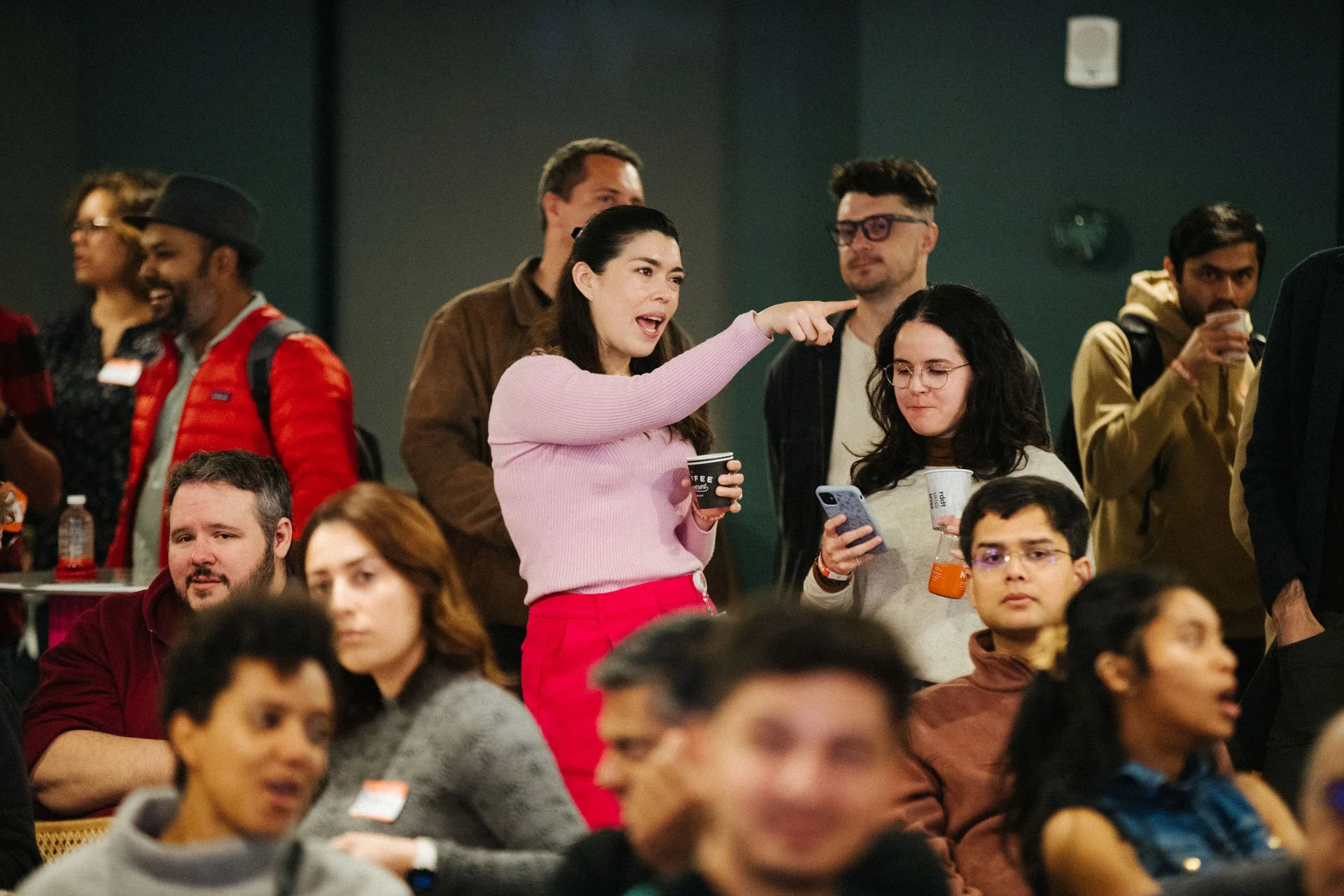 A group of diverse people at a crowded event, with a woman in a pink sweater and red pants gesturing as she talks, holding a cup, while others around her are listening, drinking, or looking at their phones.