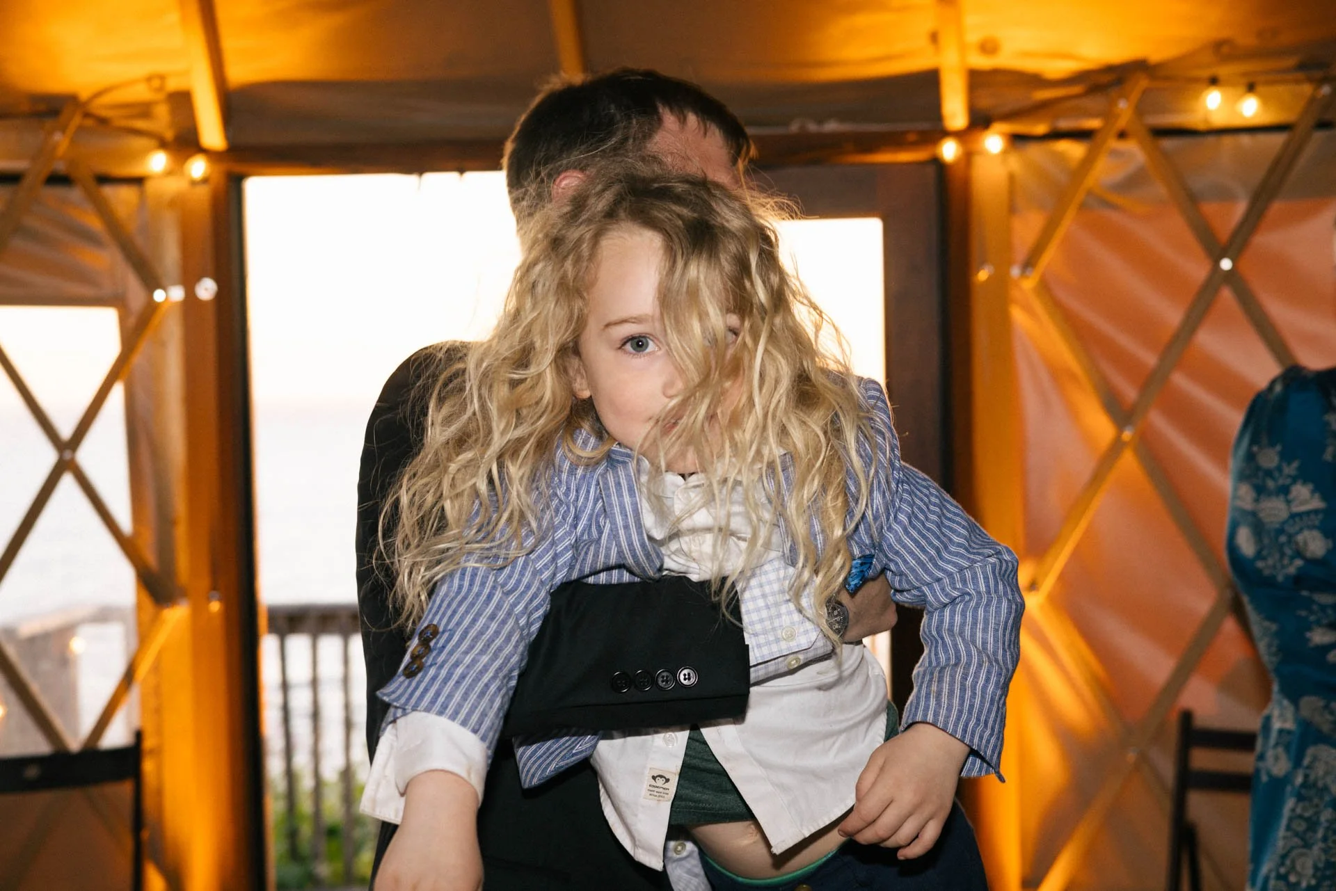 A young girl with long curly blonde hair being carried by an adult inside a wooden yurt with glowing string lights.