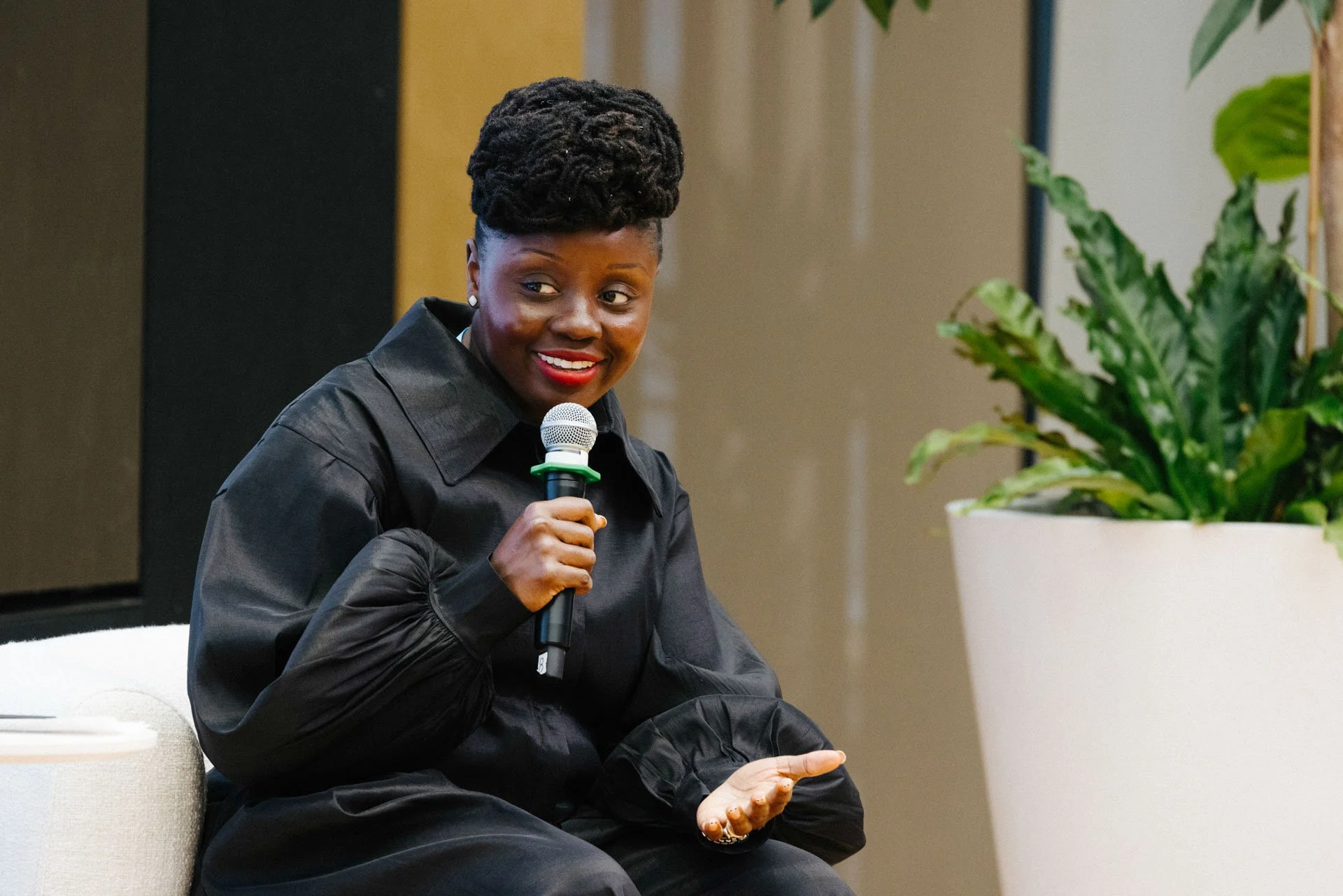 A woman sitting and smiling while holding a microphone, wearing a black jacket, next to a large potted green plant.