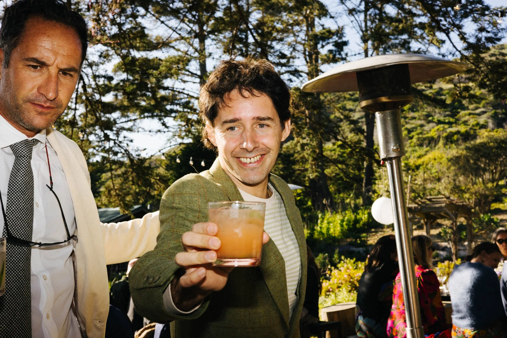 A smiling man in a light green blazer holding a glass of drink outdoors at a gathering, with a lighting heater nearby and other people seated in the background surrounded by trees.