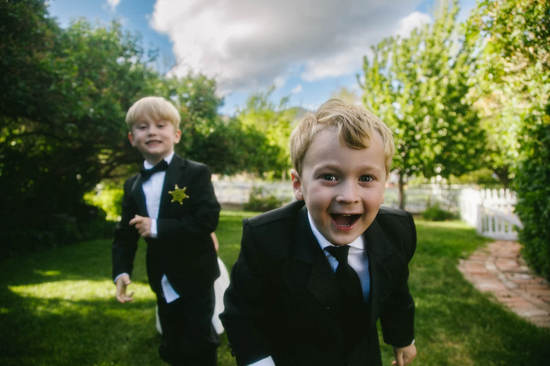 Two young boys dressed in tuxedos and police badges, playing outside on a sunny day in a grassy garden with trees and a brick pathway.