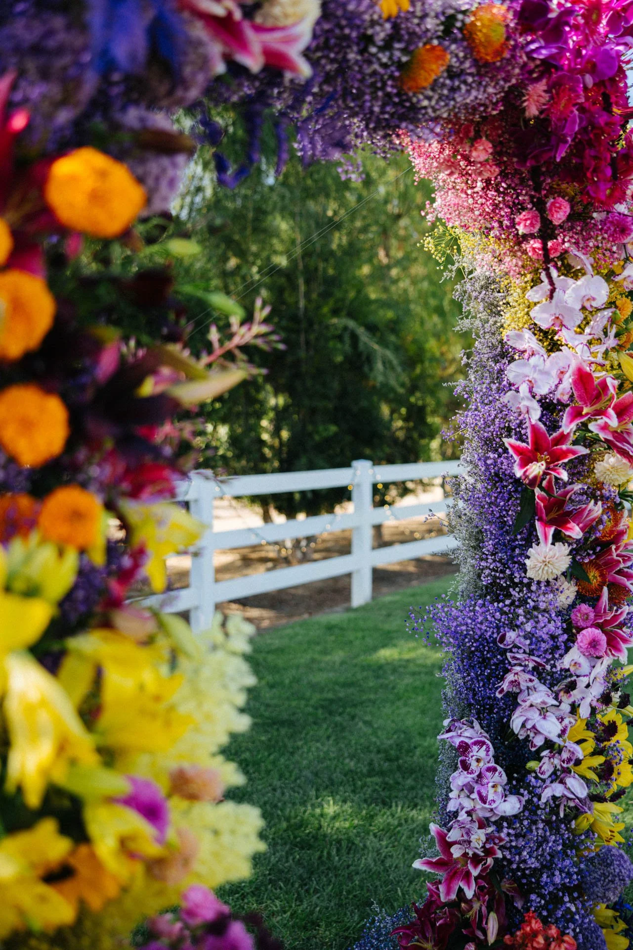 Colorful floral archway with purple, pink, yellow, and white flowers, set outdoors with green grass and a white fence in the background.