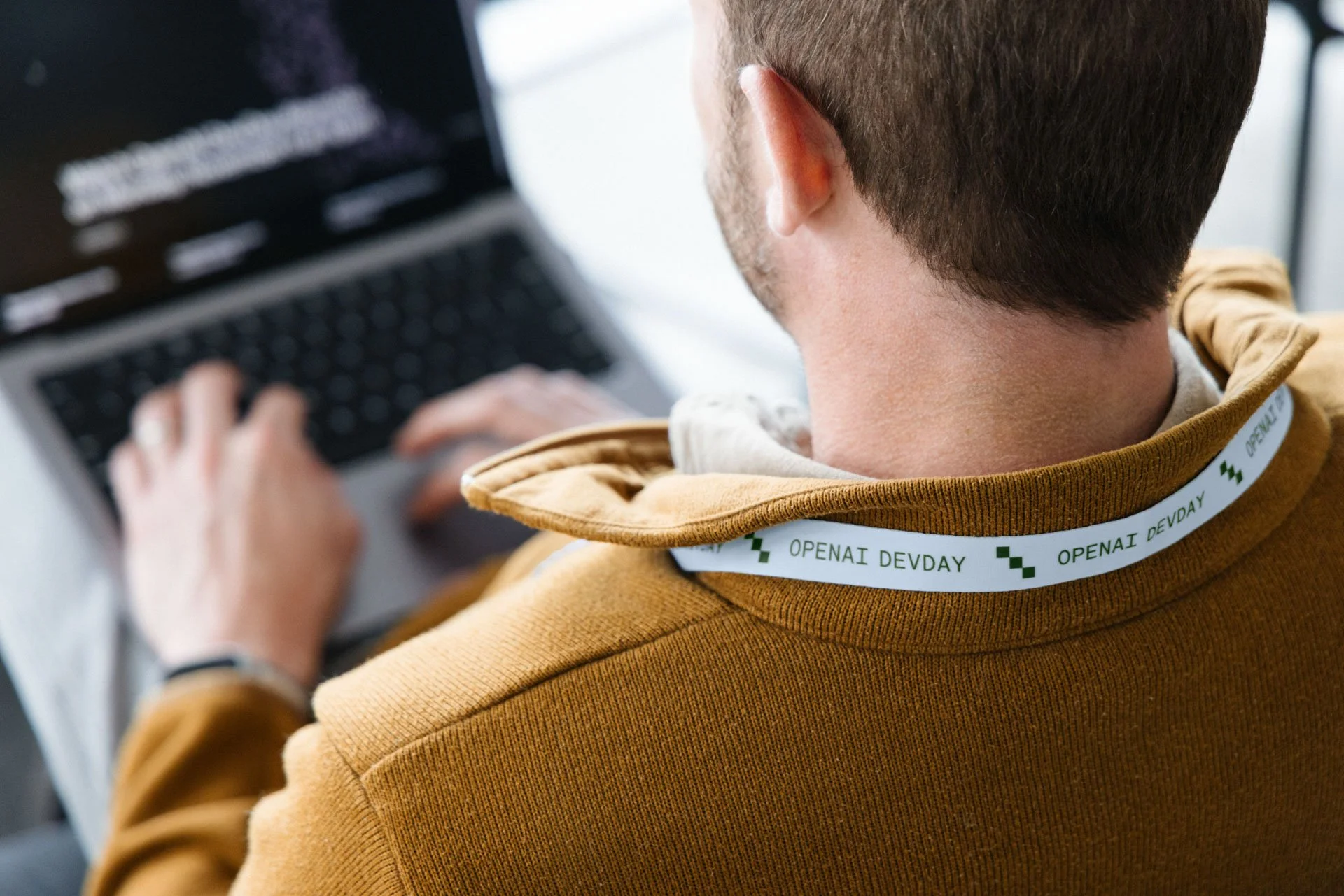 A man with short brown hair wearing a mustard yellow hoodie, viewed from behind, working on a laptop.