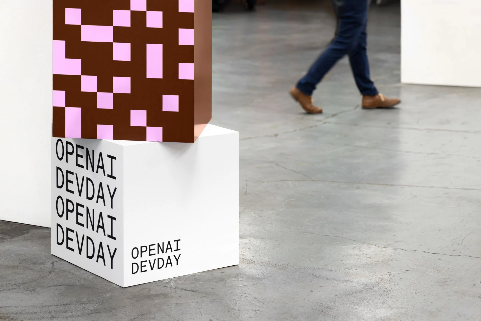 A white display pedestal with the words 'OPENAI DEVDAY' printed on its sides in black text. A colorful geometric cube with pink and brown pattern is placed on top of the pedestal. In the background, a person's legs are visible walking away.