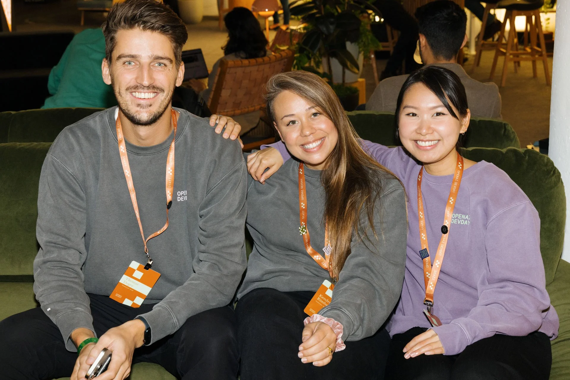 Three young adults sitting on a green sofa at an indoor event, smiling at the camera, wearing casual clothing with event badges around their necks.