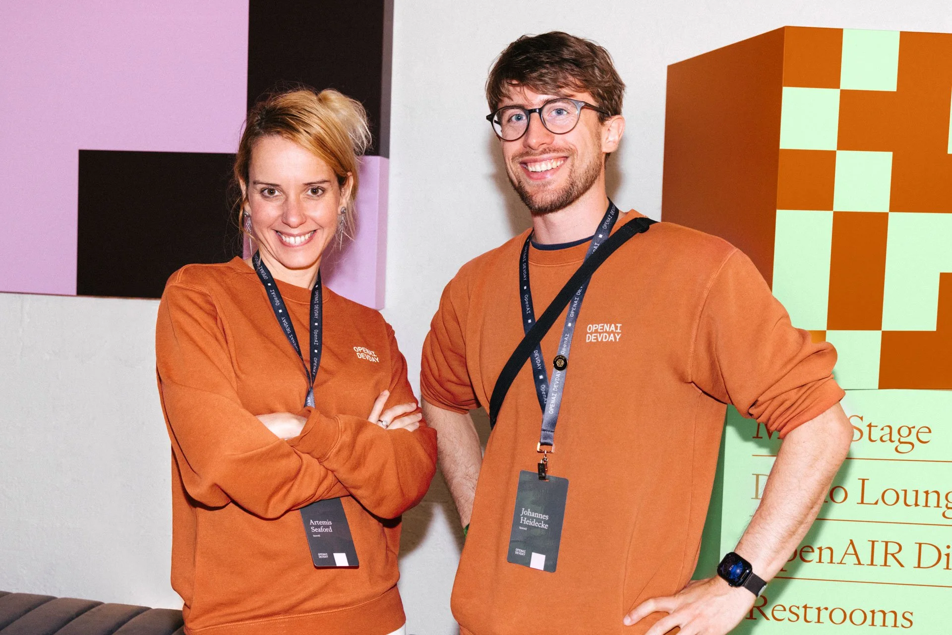 Two smiling conference attendees, a woman and a man, standing indoors with a colorful background and a sign. They are wearing orange sweatshirts and conference badges.