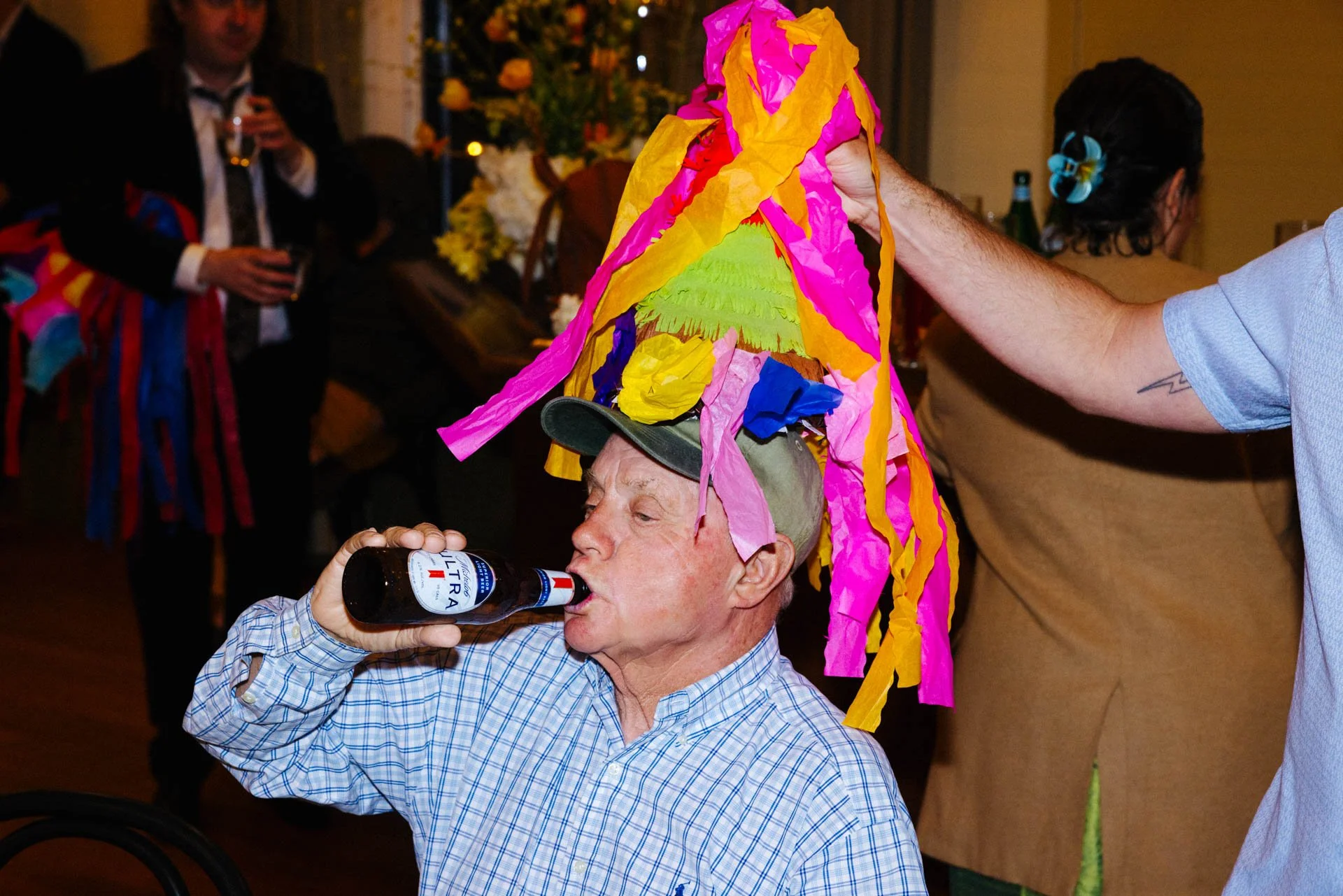 An elderly man wearing a colorful paper hat and baseball cap, drinking a bottle of Budweiser beer at a party, with other guests in the background.