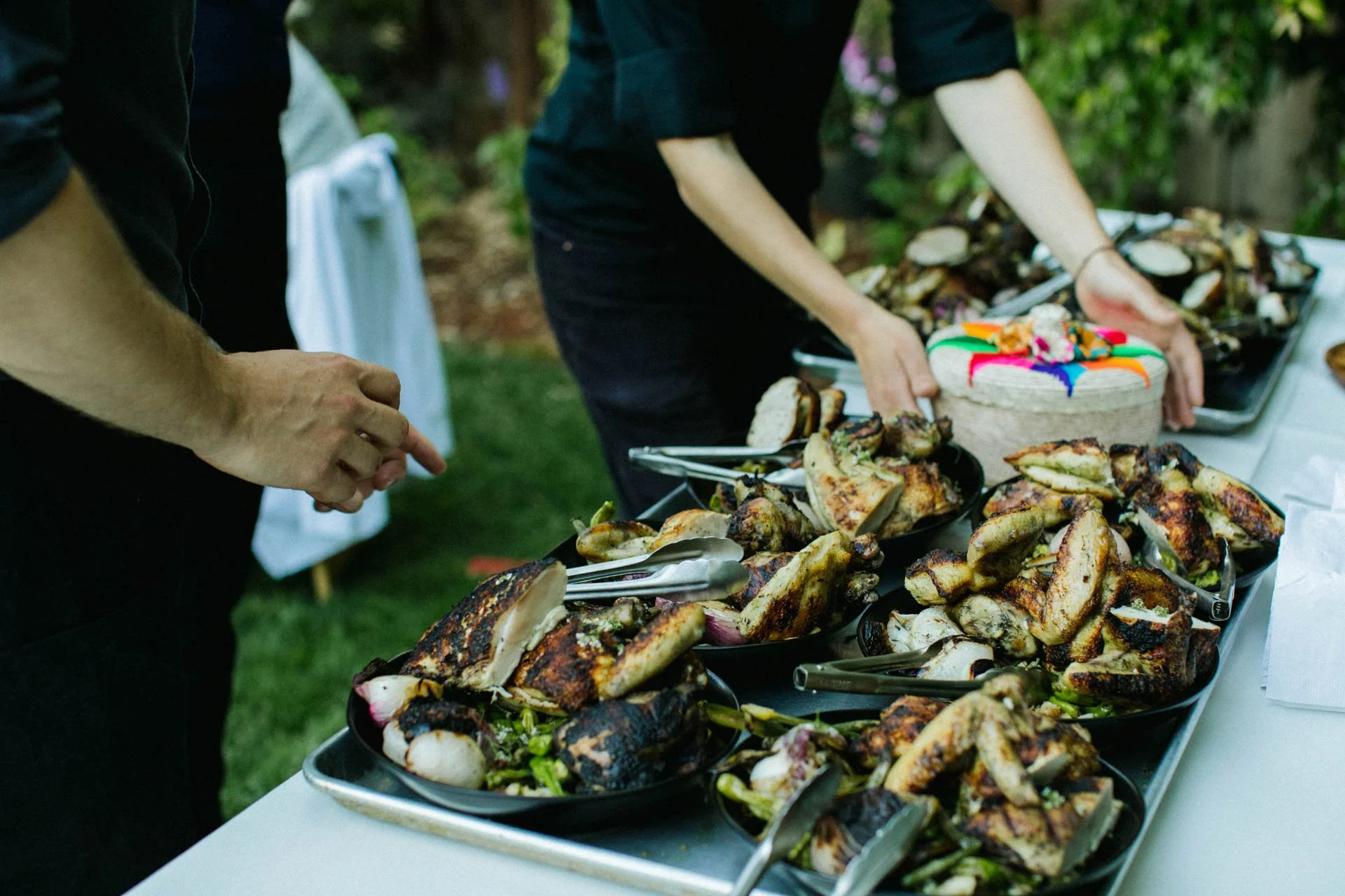 People serving grilled chicken and vegetables at an outdoor event with a decorated cake on the table.