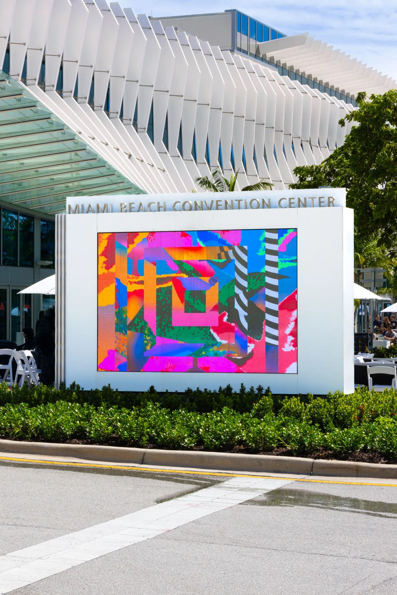 Colorful digital art display at Miami Beach Convention Center exterior, with modern architectural design, white structural elements, and green landscaped bushes in foreground. Photography of Adobe Max conference in Miami for Play.Studio.  