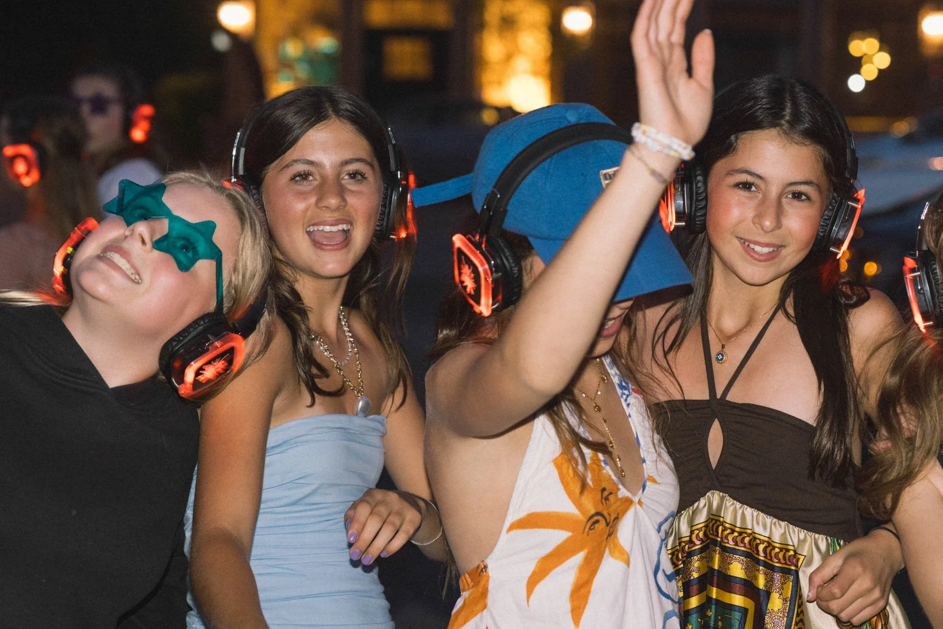 Group of young women at a nighttime outdoor event, wearing headphones and enjoying music, some dancing or smiling.