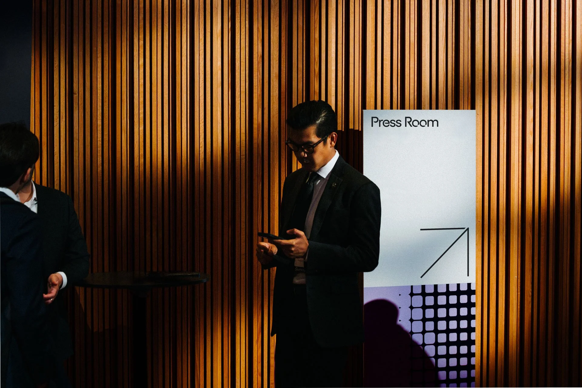 A man in a suit and glasses looking at his phone, standing in front of a wooden wall with a sign that reads 'Press Room' during a formal event.