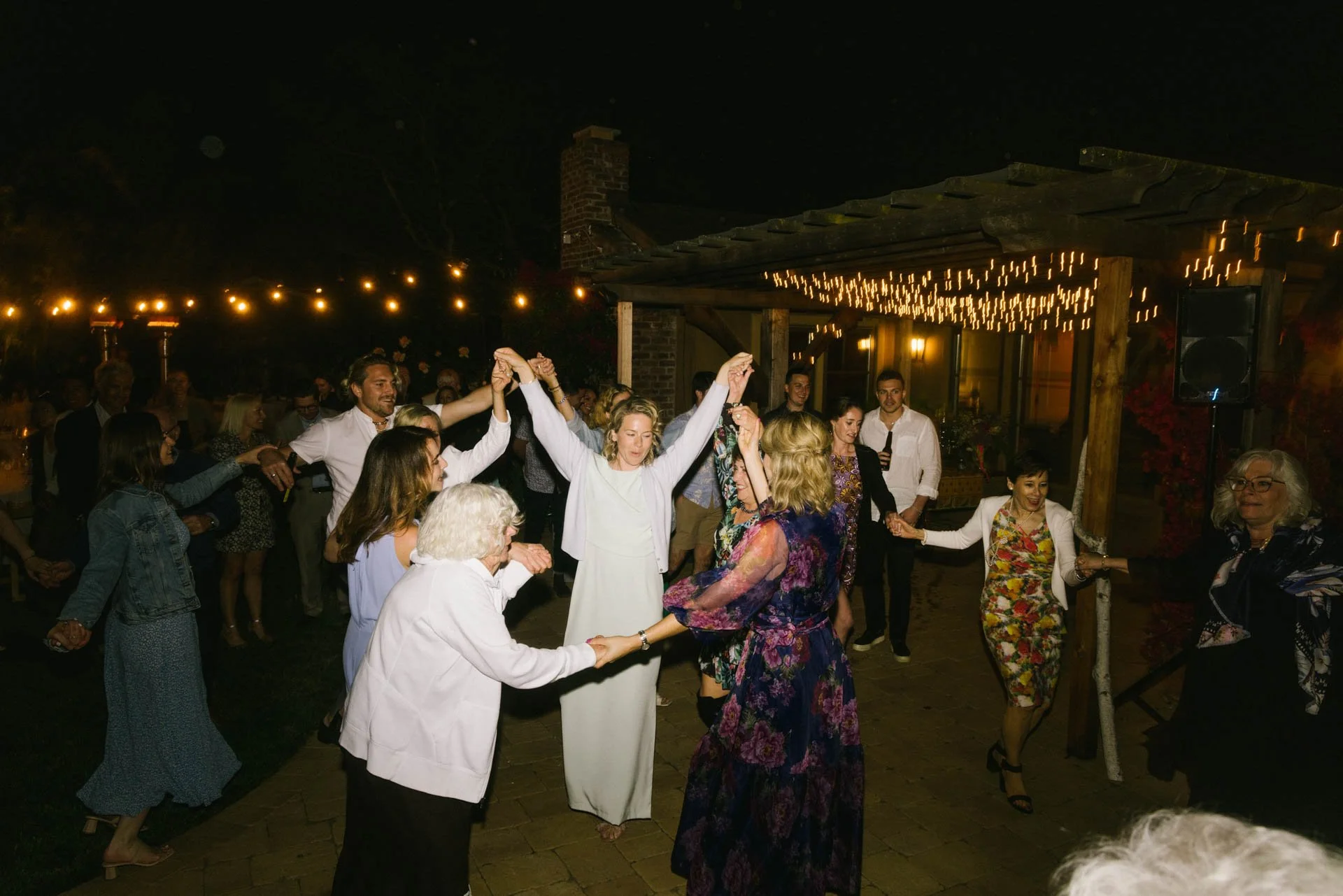 People dancing and celebrating at an outdoor night event with string lights in background.