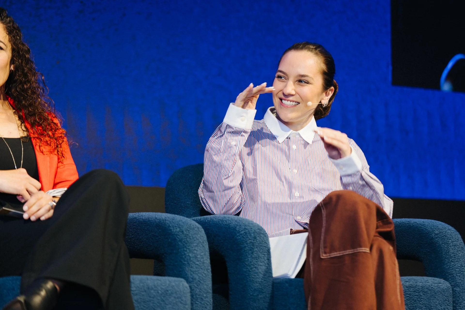 Woman smiling and gesturing with hands while sitting on a blue armchair at a panel discussion or conference.