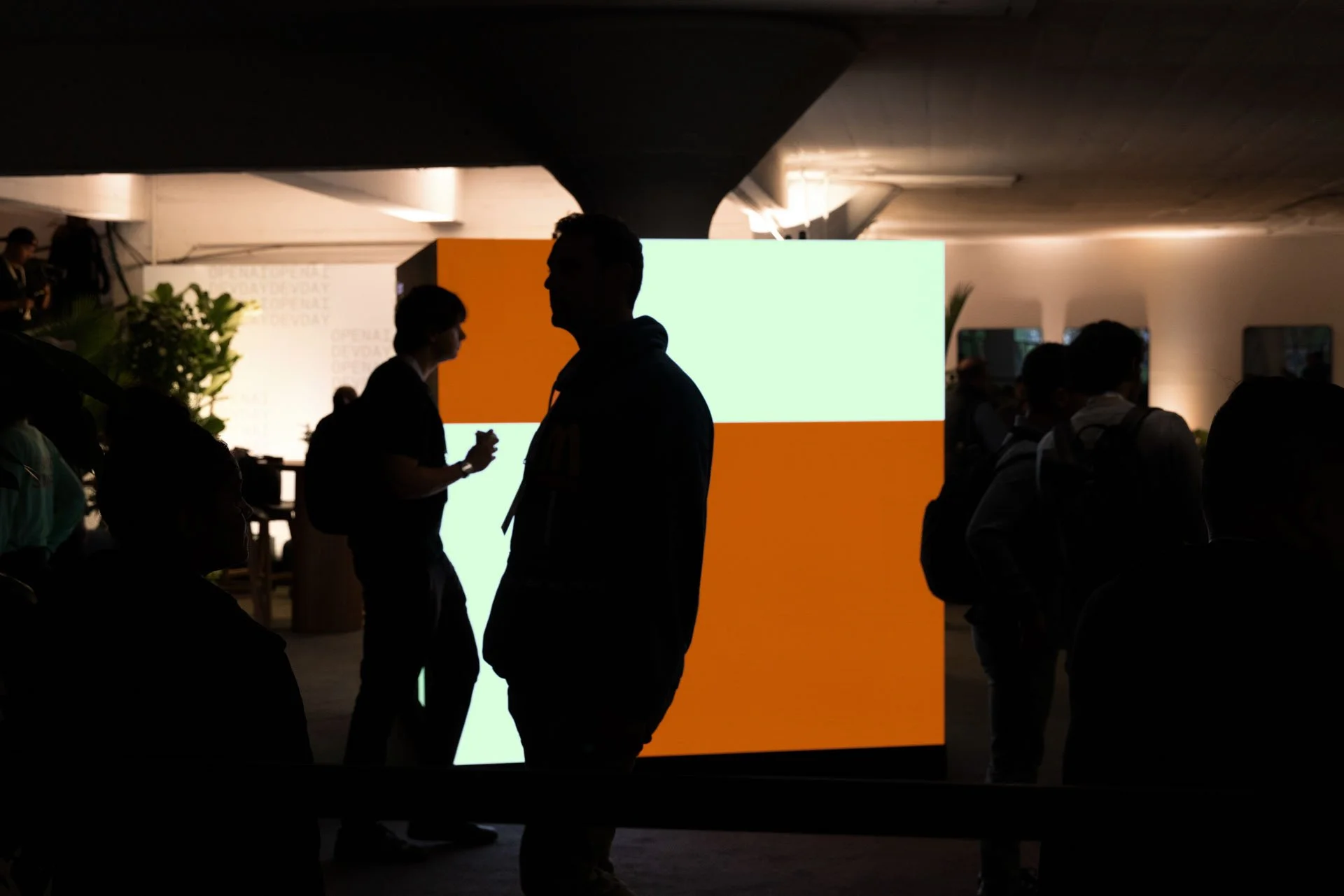 Silhouettes of people standing and talking in front of a brightly lit, colorful display or sign.