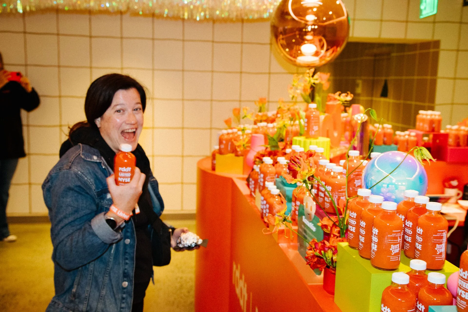 A woman with black hair smiling and holding a bottle of orange juice, standing in front of a colorful display of bottles of orange juice, flowers, and decorative items at an event.