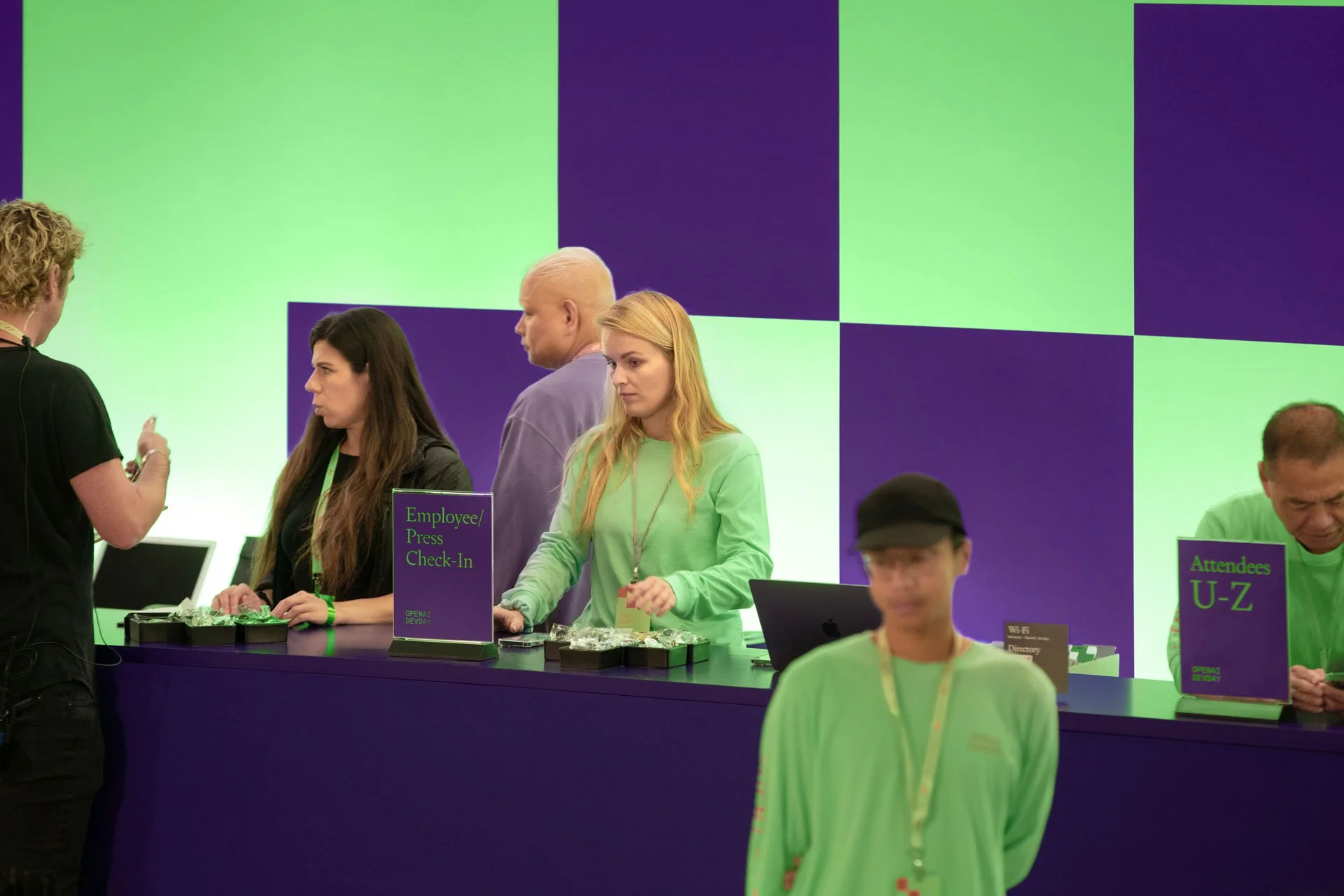 Conference registration area with staff at check-in desk, signs indicating employee and attendee registration, and a large colorful digital screen behind the staff.