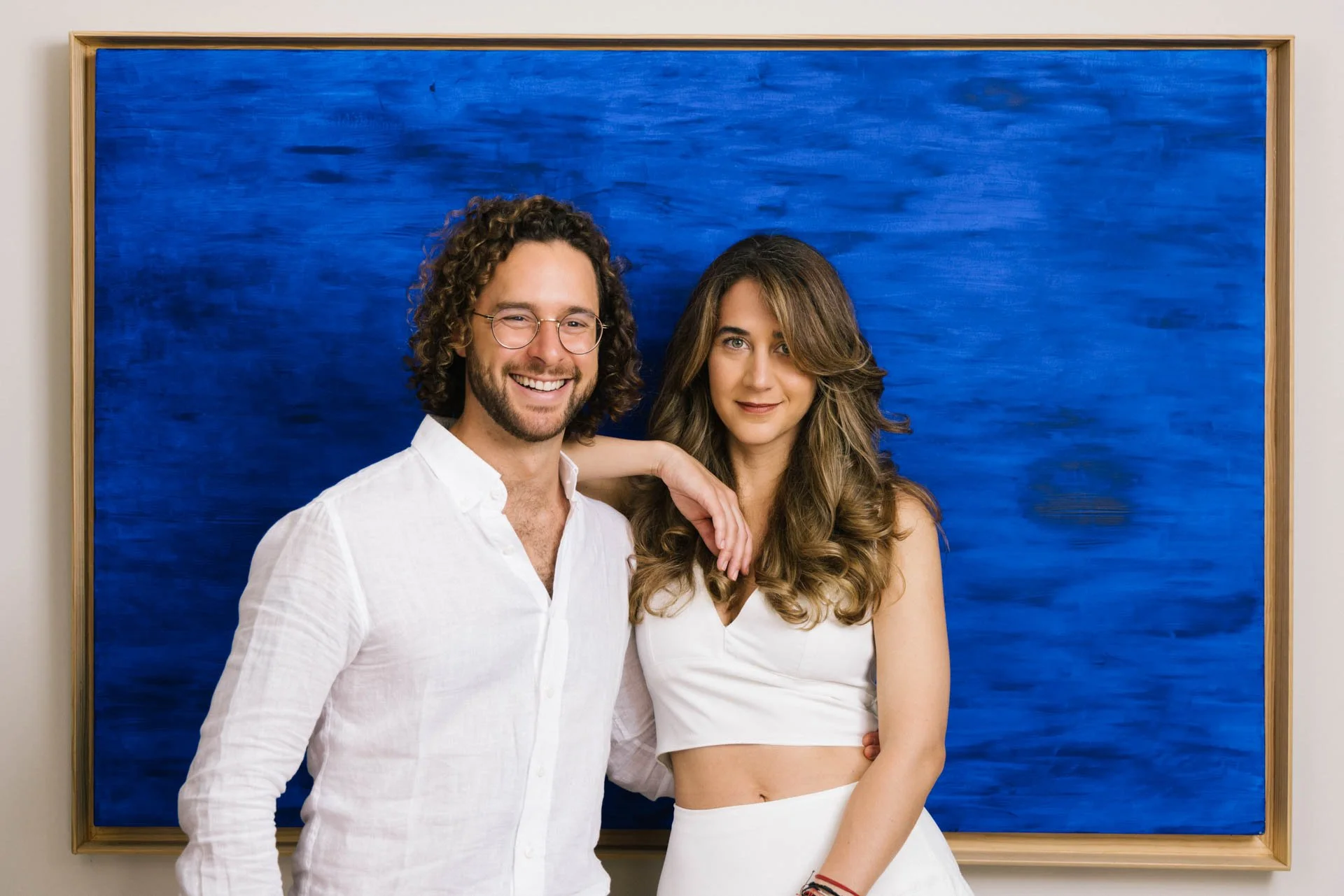 A man and a woman standing in front of a large blue abstract painting, smiling at the camera. The man has curly hair, glasses, and a beard, wearing a white shirt. The woman has long brown hair, wearing a white crop top and a white skirt, with her han