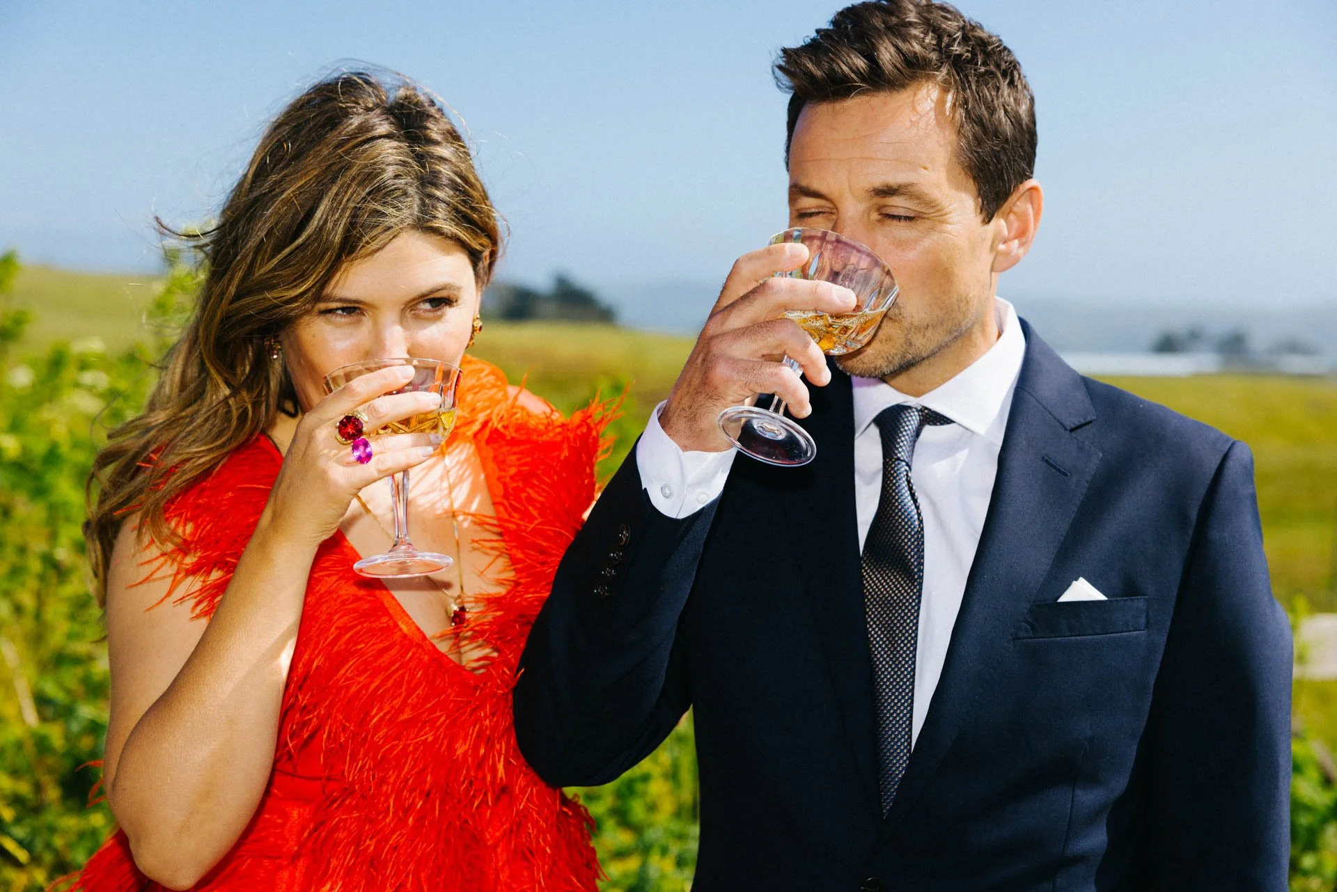 A man and a woman dressed formally are outdoors, drinking from glasses of wine, with a green landscape and cloudy sky in the background.