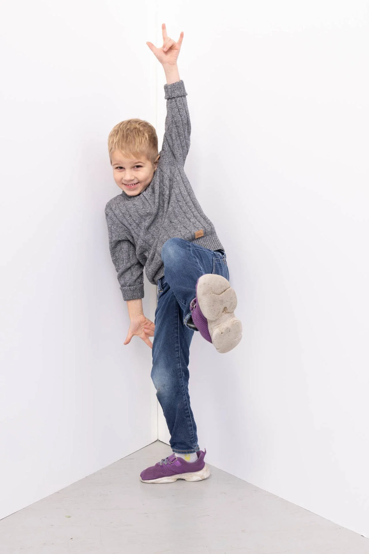 Young boy smiling, one arm raised, standing against white walls, lifting one foot to show the sole of his purple sneaker.
