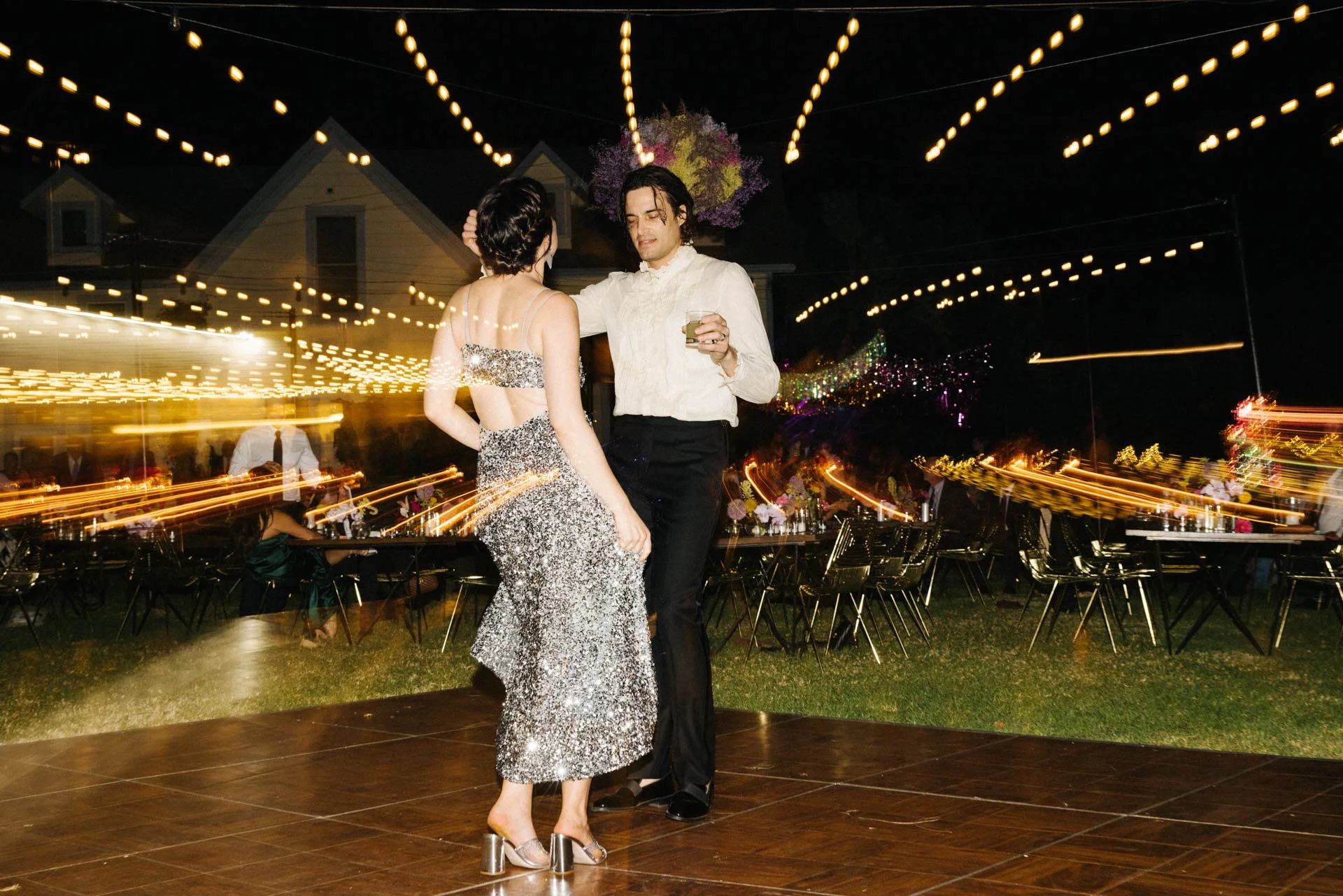 Two people dancing at a nighttime event with string lights overhead, tables and chairs in the background, one holding a drink, the other wearing a sparkly dress, with a blurred motion effect.