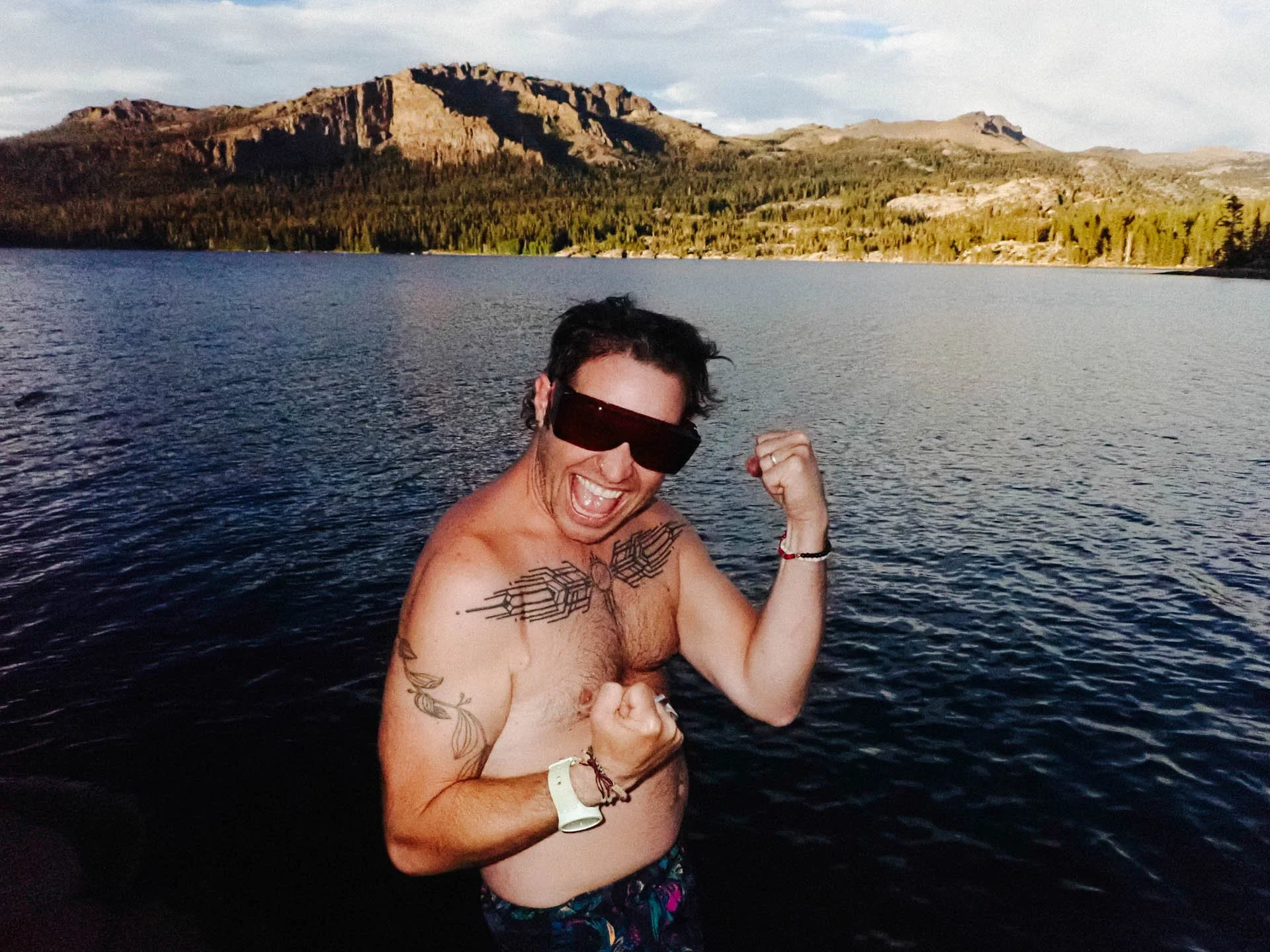 A cheerful man with tattoos wearing dark sunglasses, shirtless and flexing his arm, standing near a large body of water with mountains and a forest in the background.