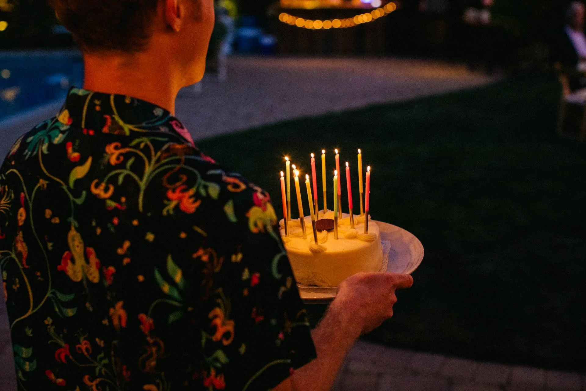 Person holding a birthday cake with lit candles at an outdoor evening celebration.