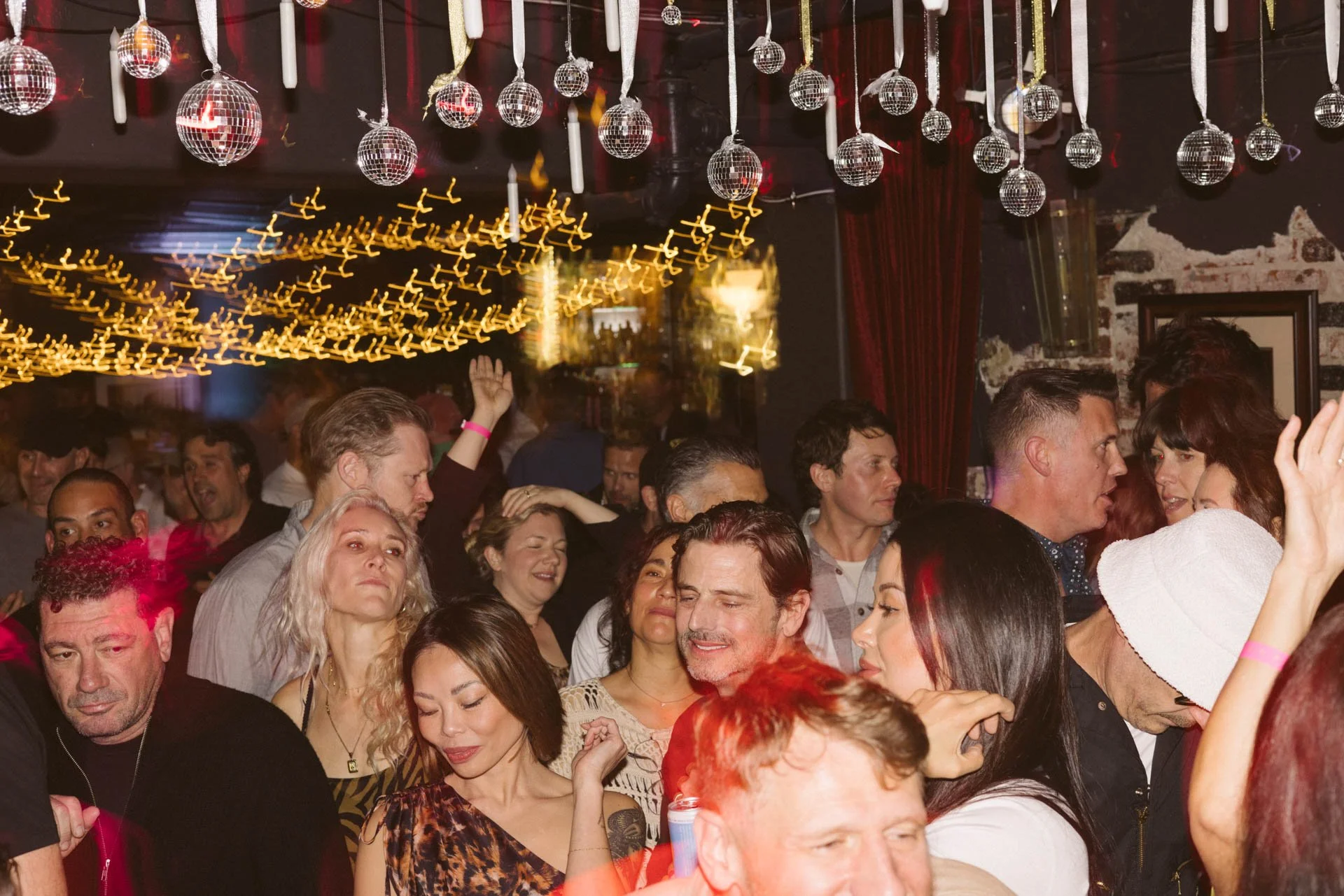 Crowd of people dancing and socializing at a party or nightclub with hanging disco balls and string lights.