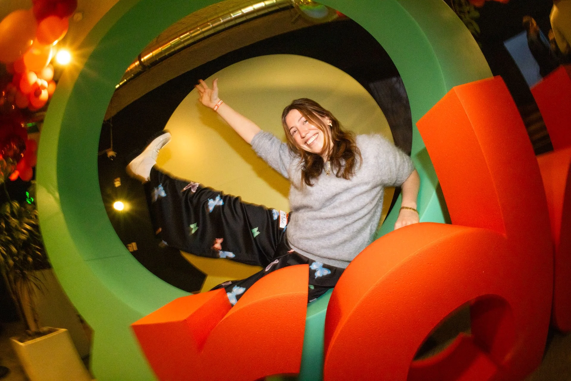 A woman sitting inside a colorful, circular sculpture, smiling and making a peace sign with her right hand.